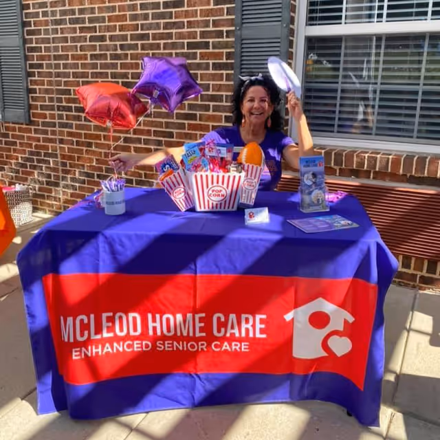 A smiling woman sits at an outdoor table with a McLeod Home Care banner.