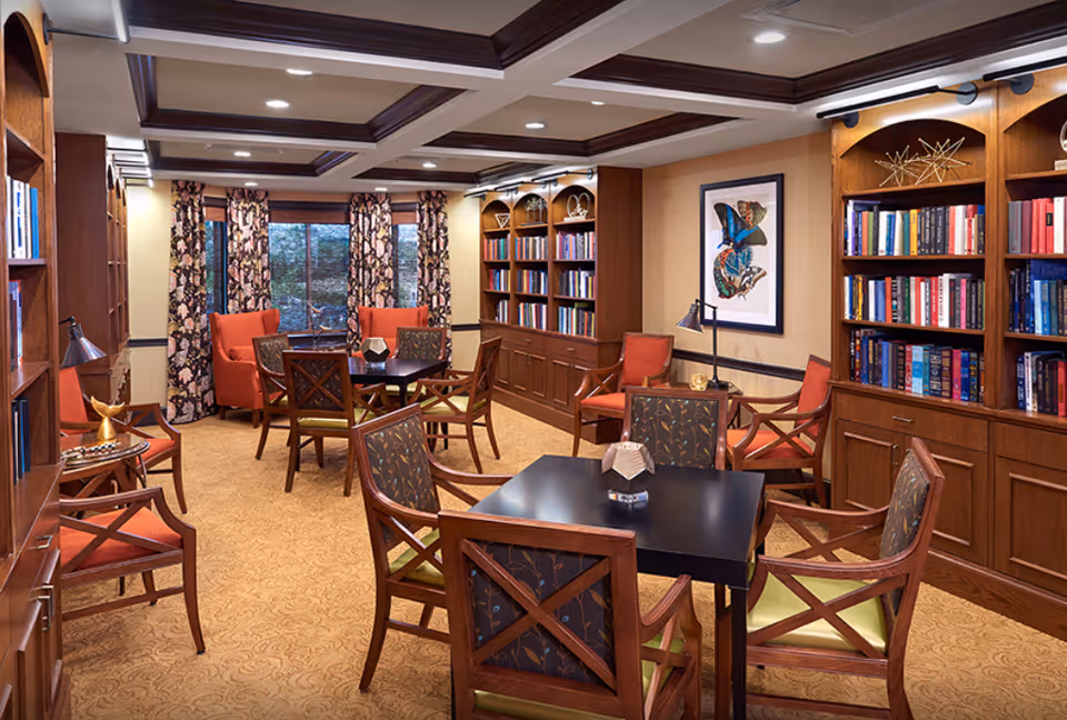 Cozy senior living common room with wooden bookshelves, multiple tables and chairs, patterned curtains and armchairs.