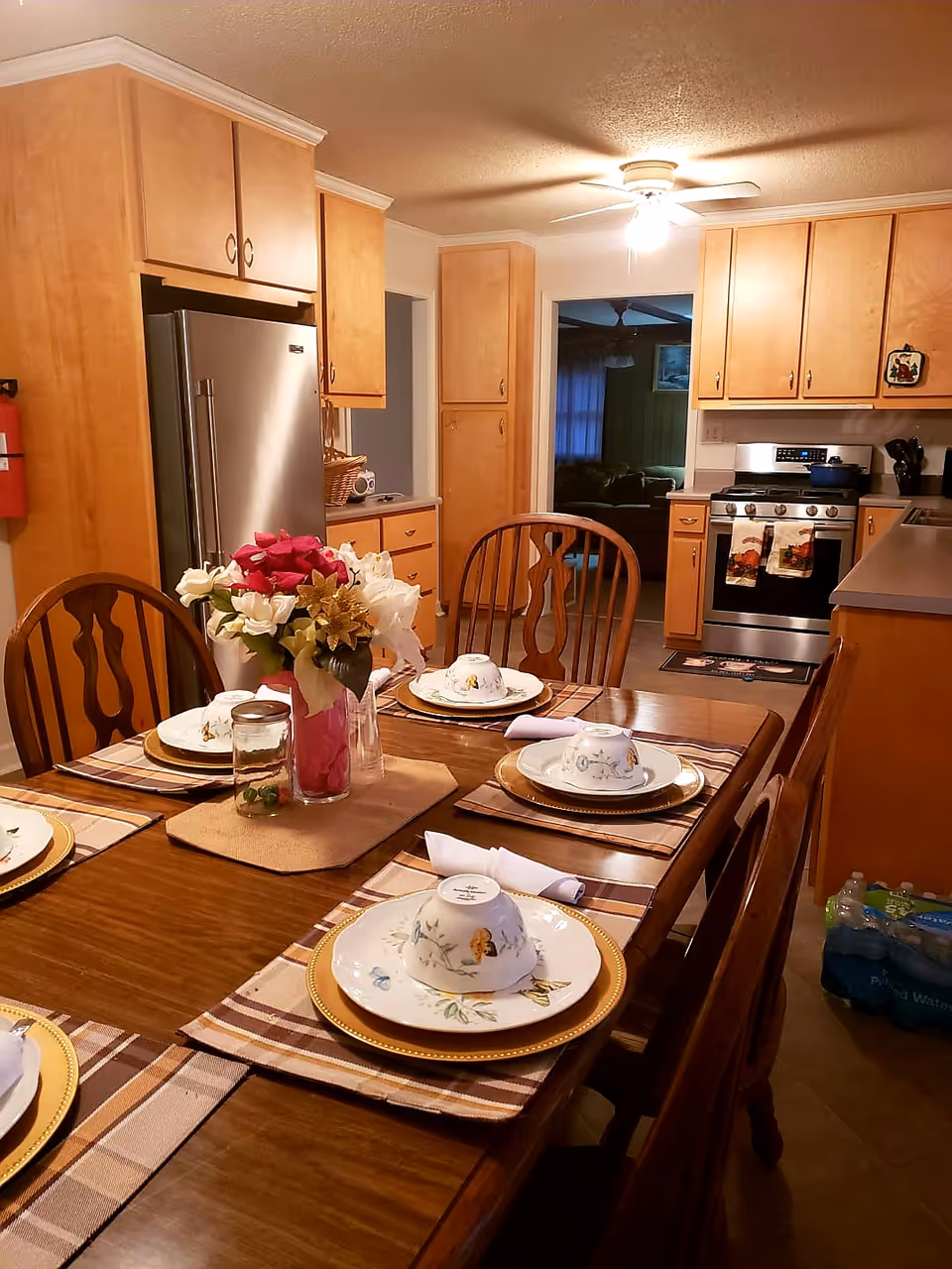A cozy kitchen and dining area with wooden cabinets and a stainless steel refrigerator. A wooden dining table is set with plates, bowls, napkins, and a centerpiece of flowers. The kitchen has a stove with two decorative towels hanging on the oven door and a ceiling fan with a light above.