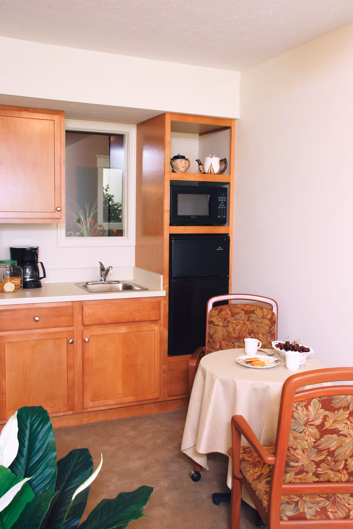 Compact kitchenette with wooden cabinets, microwave, and dining table in a senior living facility.