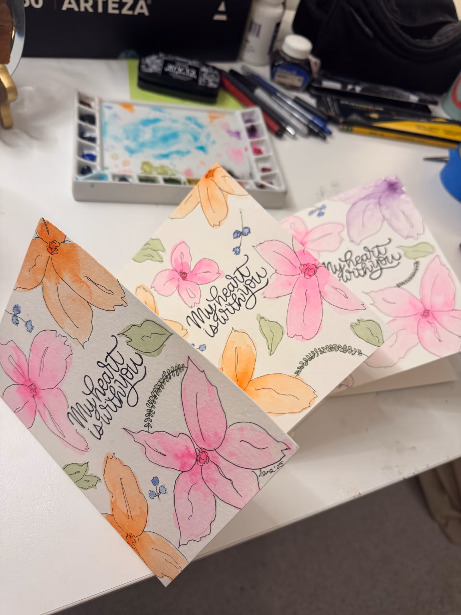 Colorful floral watercolor cards on a white craft table with painting supplies.