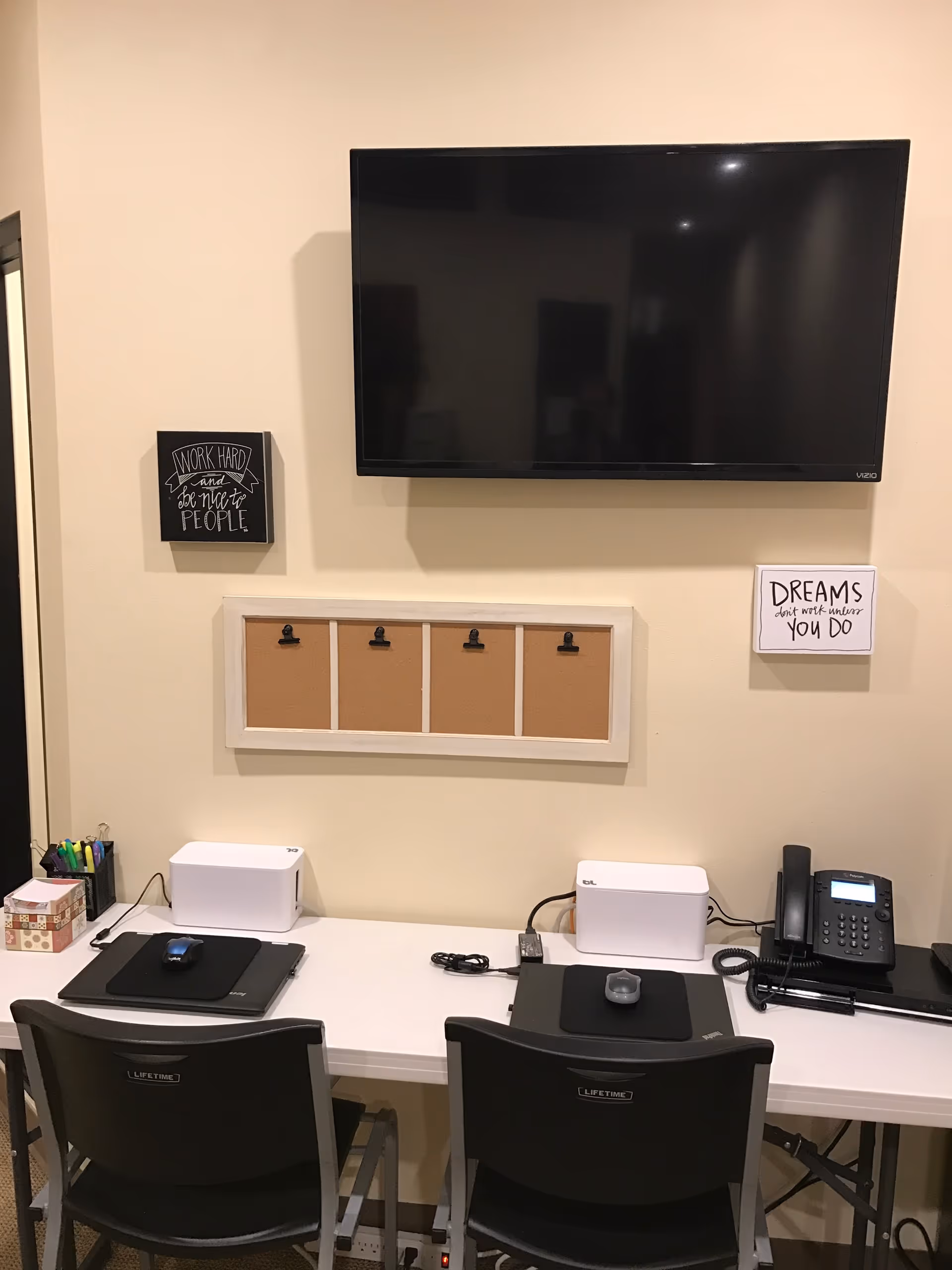 Reception desk with two workstations, mounted TV, corkboard, and office supplies.