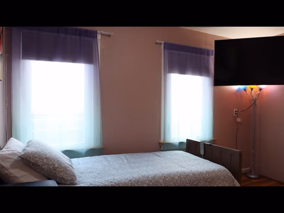 A tidy bedroom with a bed, two windows with sheer curtains, and a standing lamp.