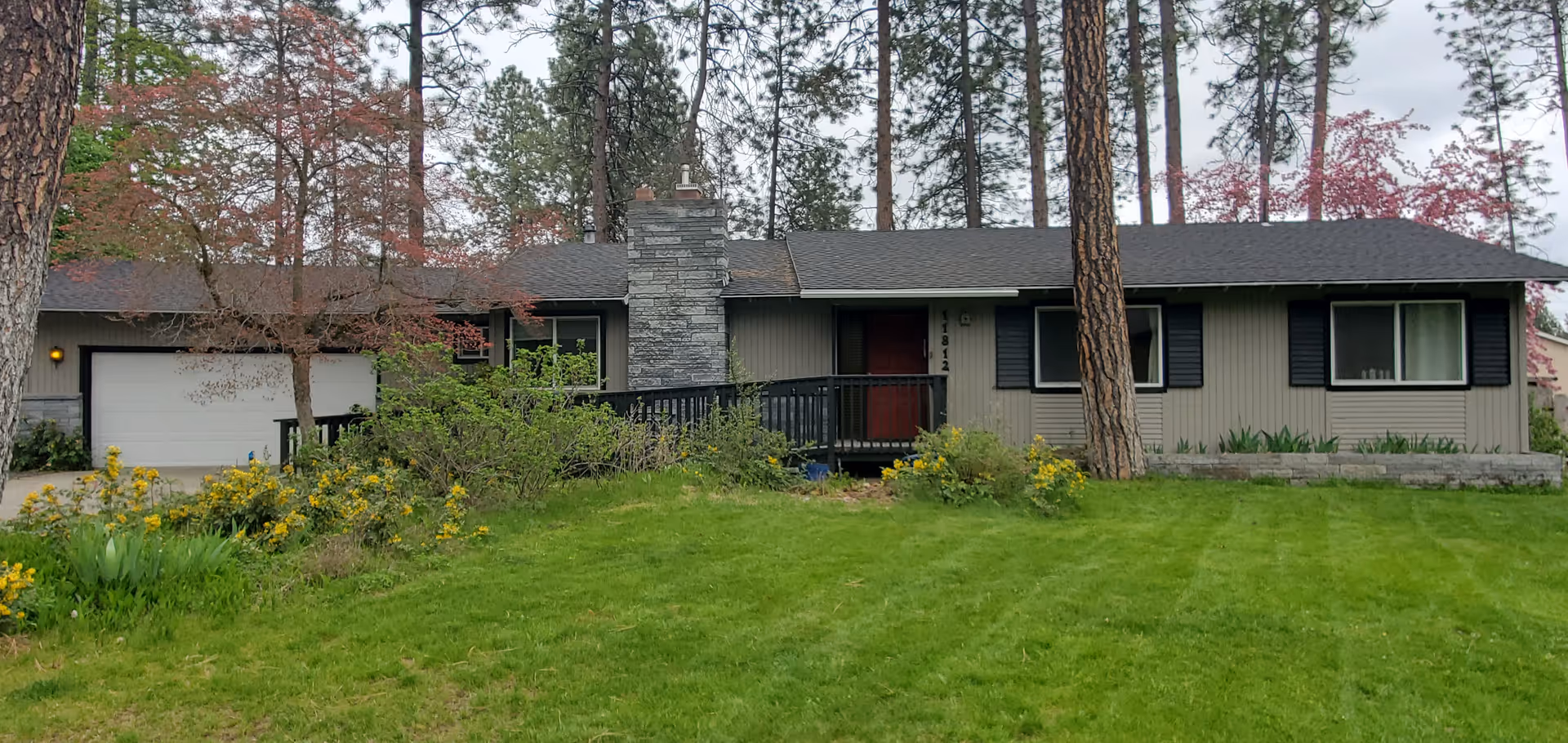 Single-story adult family home facade with front lawn, trees, and a stone chimney.