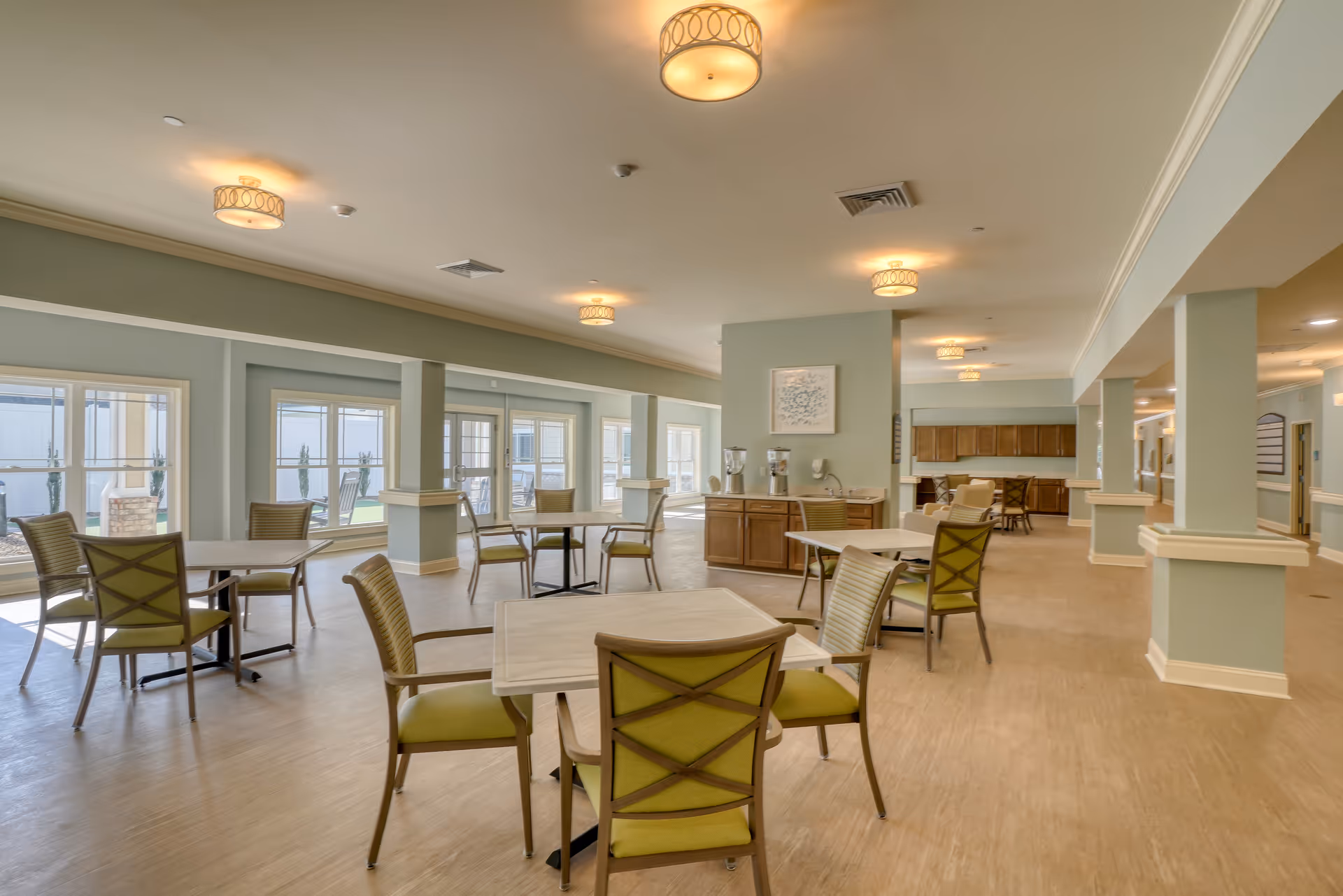 Bright dining area in Coastal Pointe Assisted Living with round tables, green chairs, and beverage station.