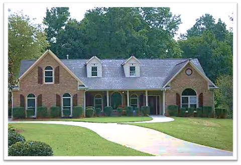 A single-story brick house with a gray shingled roof, multiple windows including dormer windows, a covered front porch with columns, and a curved driveway leading up to the entrance. The house is surrounded by a well-maintained lawn and trees in the background.