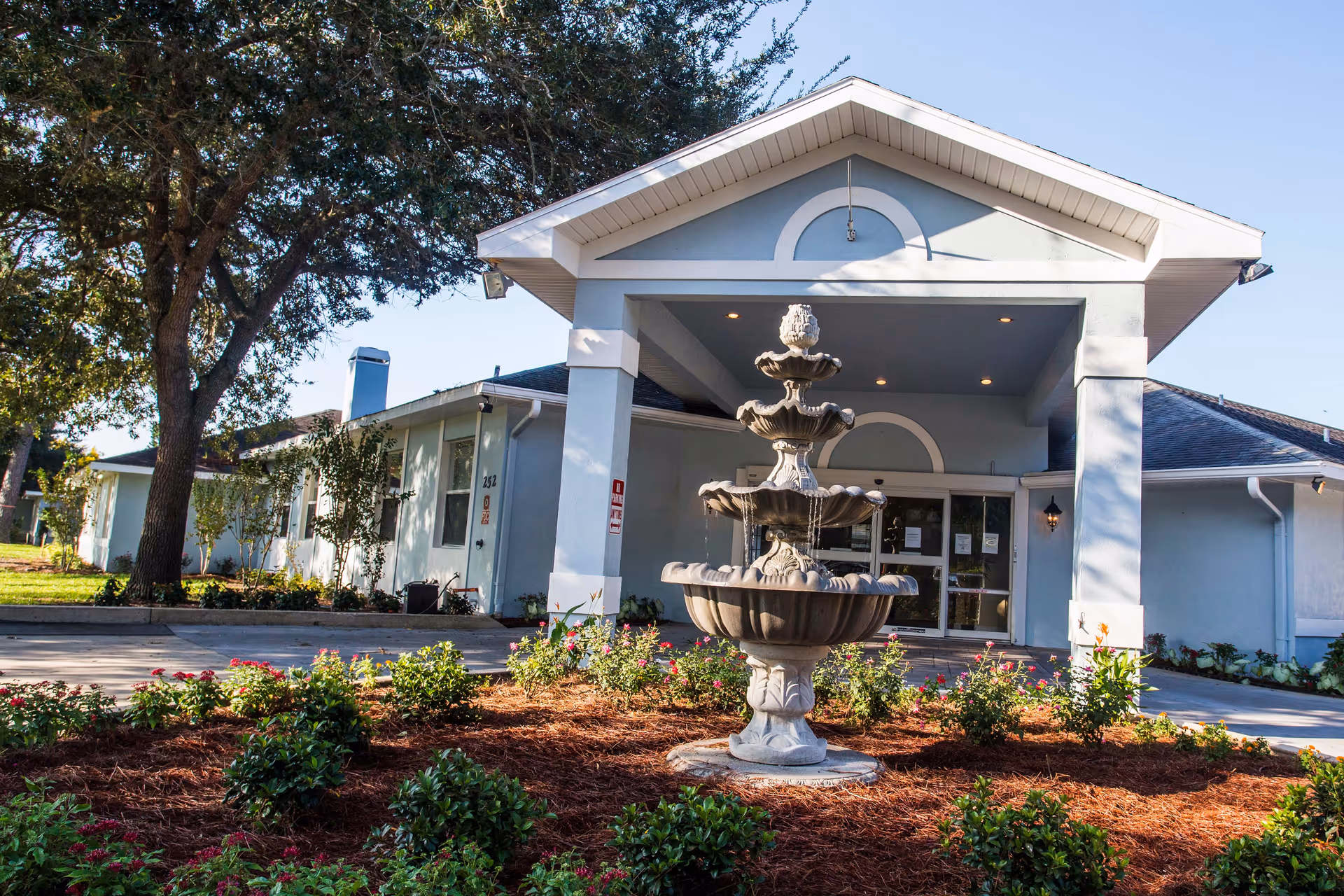 Front exterior view of Magnolia Manor Assisted Living building with a three-tiered stone fountain in the landscaped garden area in front, surrounded by small bushes and flowers, and a large tree to the left.