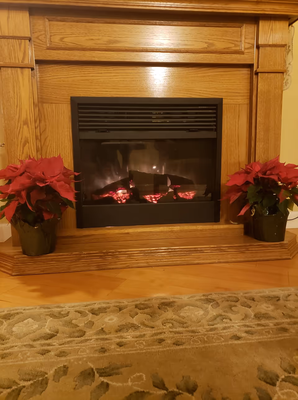 A cozy electric fireplace with a wooden mantel surrounded by two red poinsettia plants in green pots, placed on a wooden floor with a patterned beige rug in front.