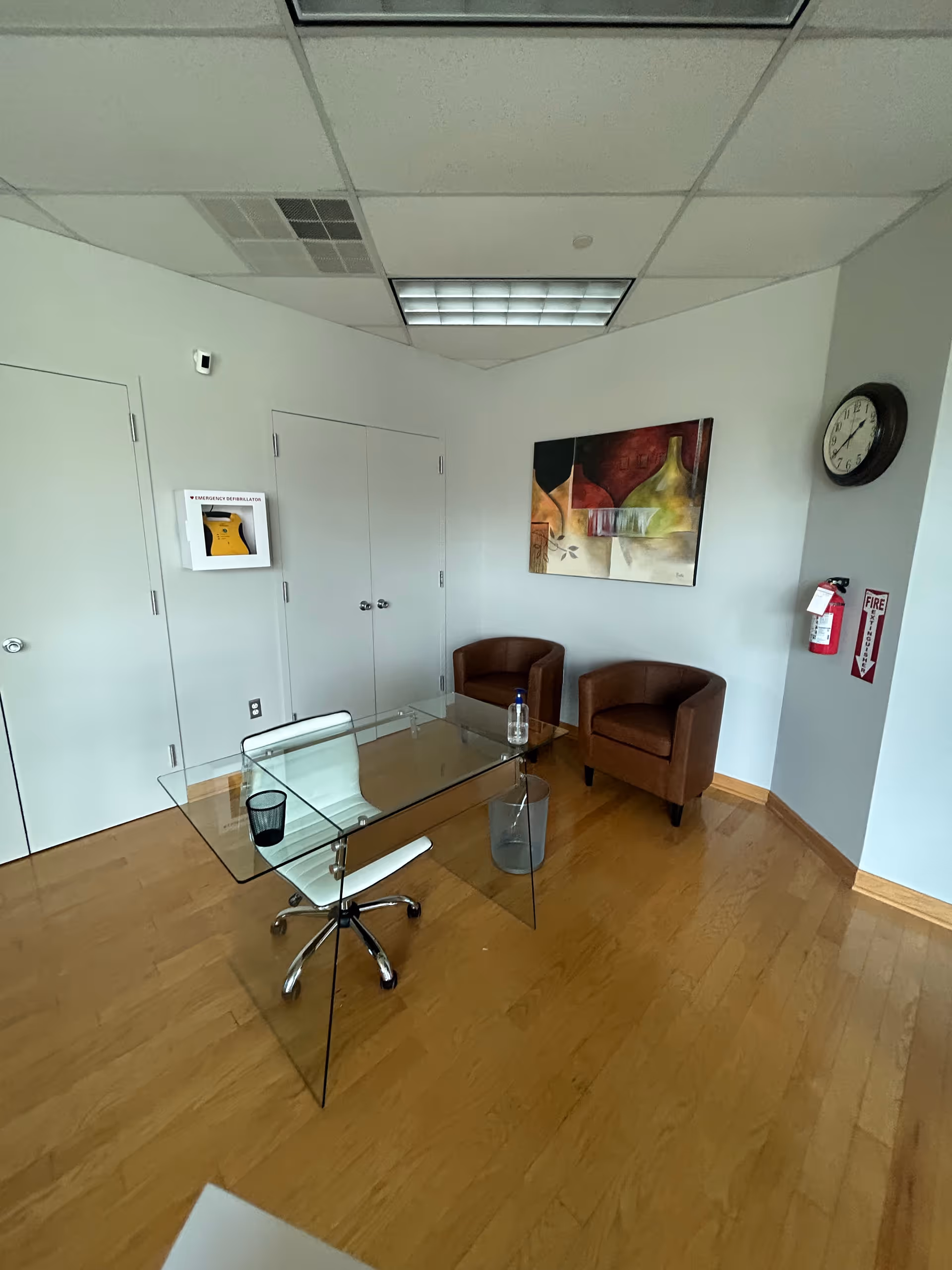 Glass desk with white chair and two brown armchairs in an office corner.