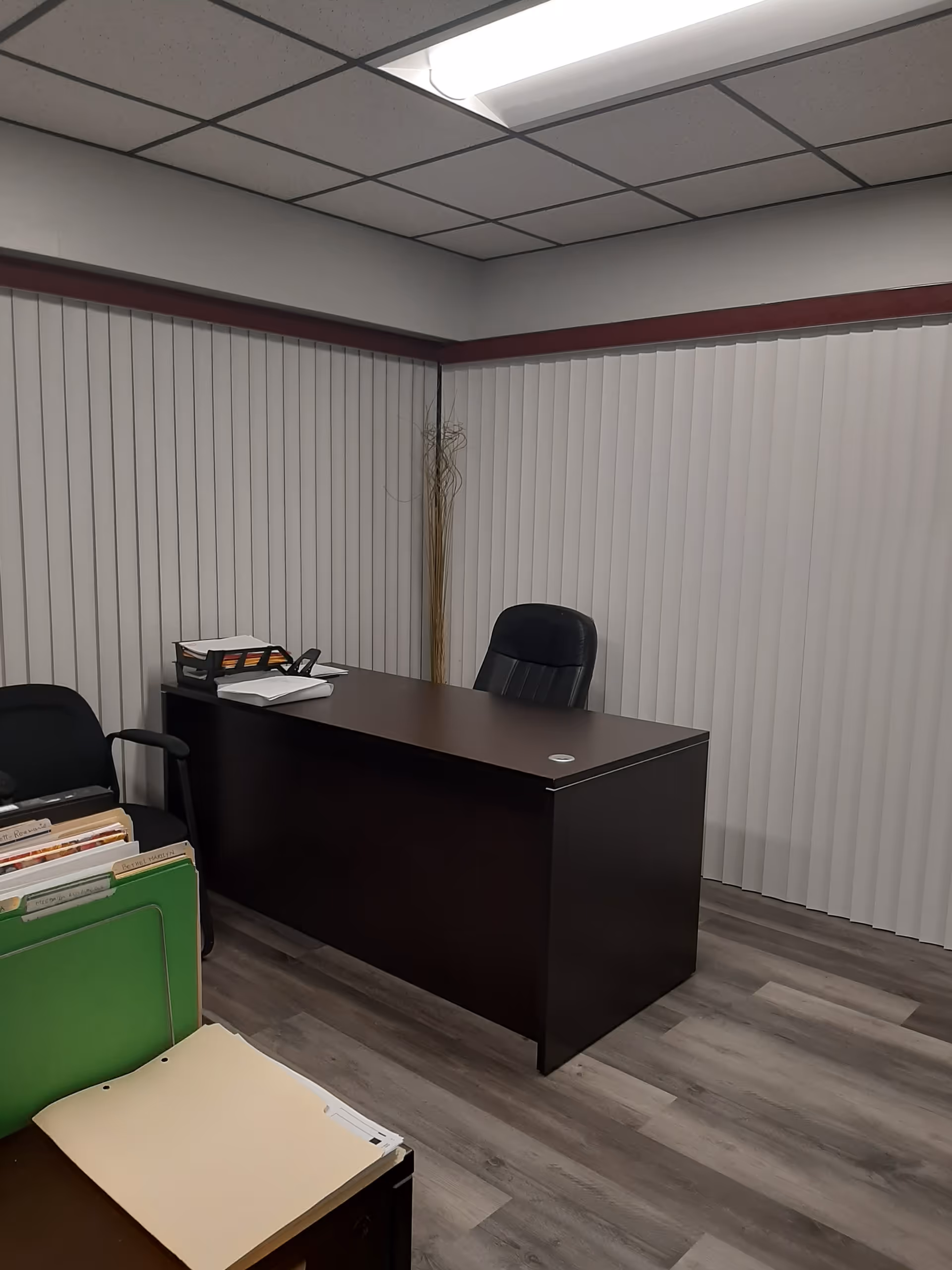 An office desk with a chair and papers in a small room with vertical blinds.