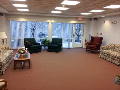Interior senior living lounge with sofas, chairs, plants, and sliding glass doors.