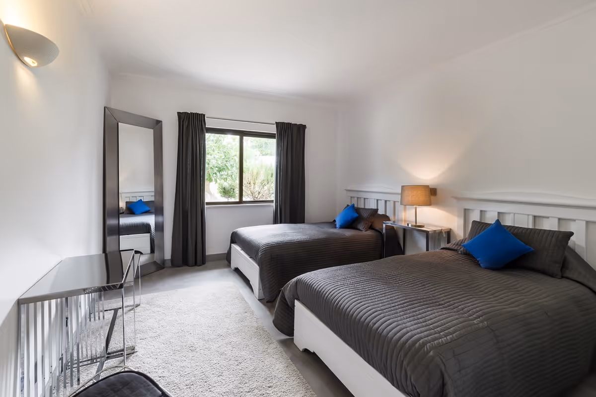 Two twin beds with gray quilts in a bright shared bedroom with a window and mirror.