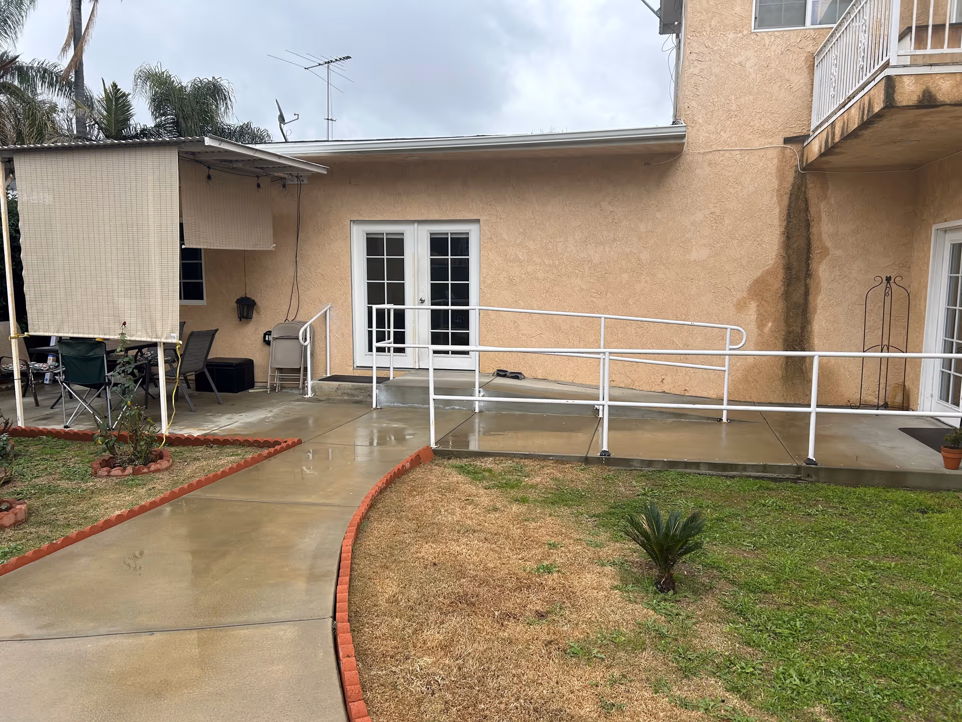 Outdoor view of a senior living facility showing a concrete pathway bordered by red bricks leading to a beige building with a white handrail ramp and double glass doors. There is a small garden area with grass and a few plants, and a covered patio area with chairs and a table on the left side.