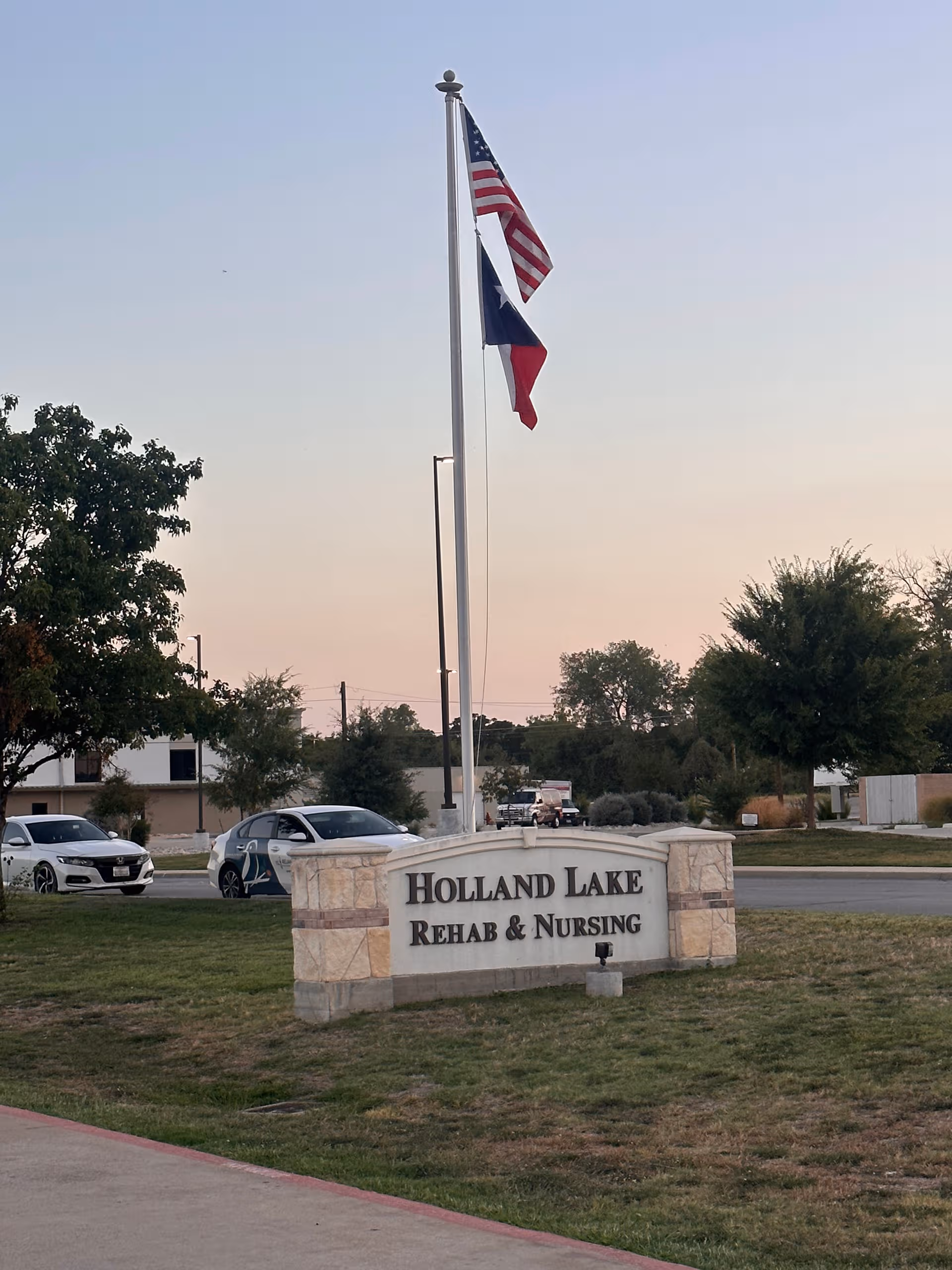 Flagpole with American and Texas flags stands near the Holland Lake Rehab and Nursing sign.