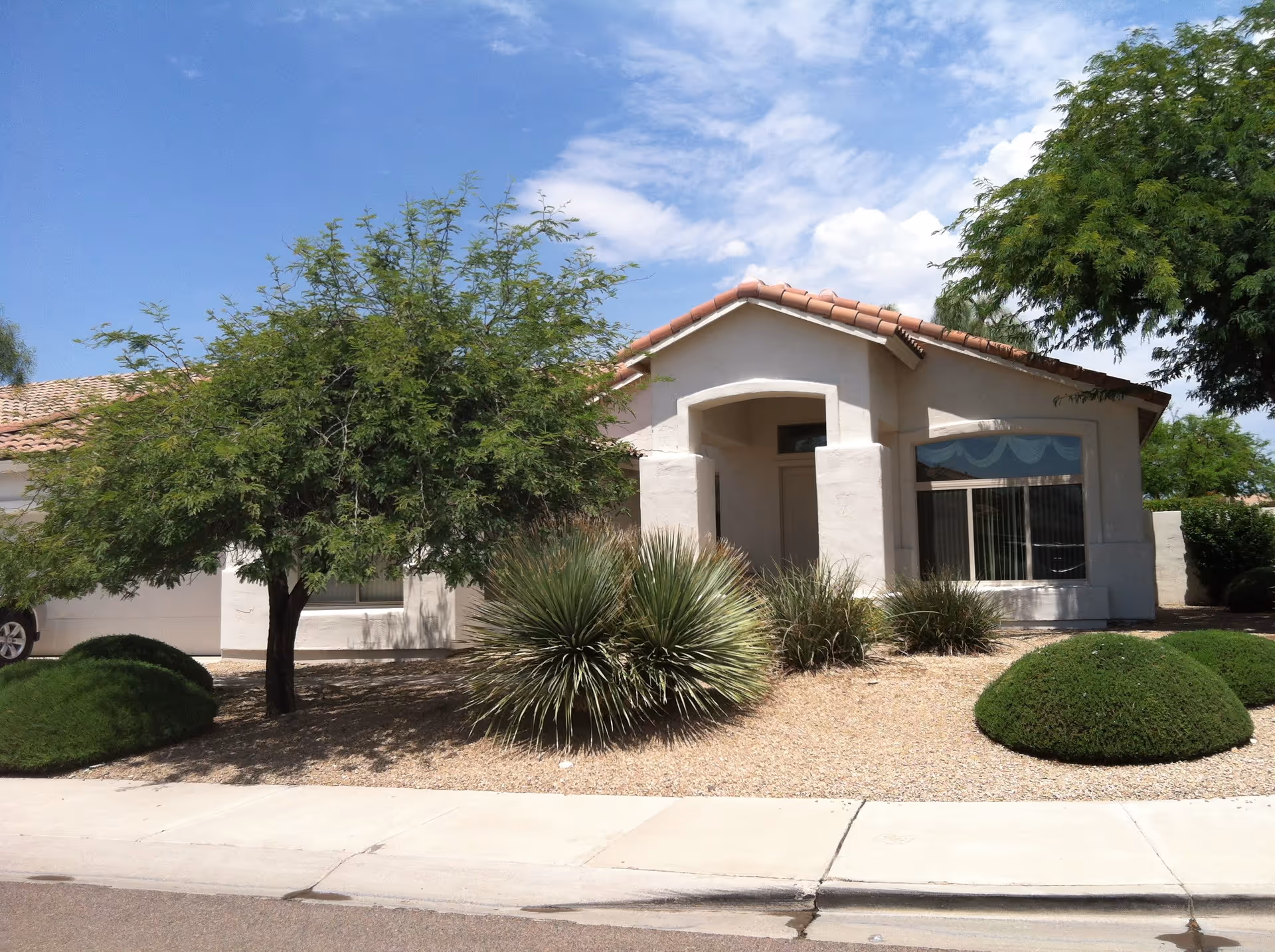 Exterior view of a single-story residential building with a tiled roof, surrounded by desert landscaping including bushes, small trees, and gravel. The building has a covered entrance and large windows with vertical blinds.