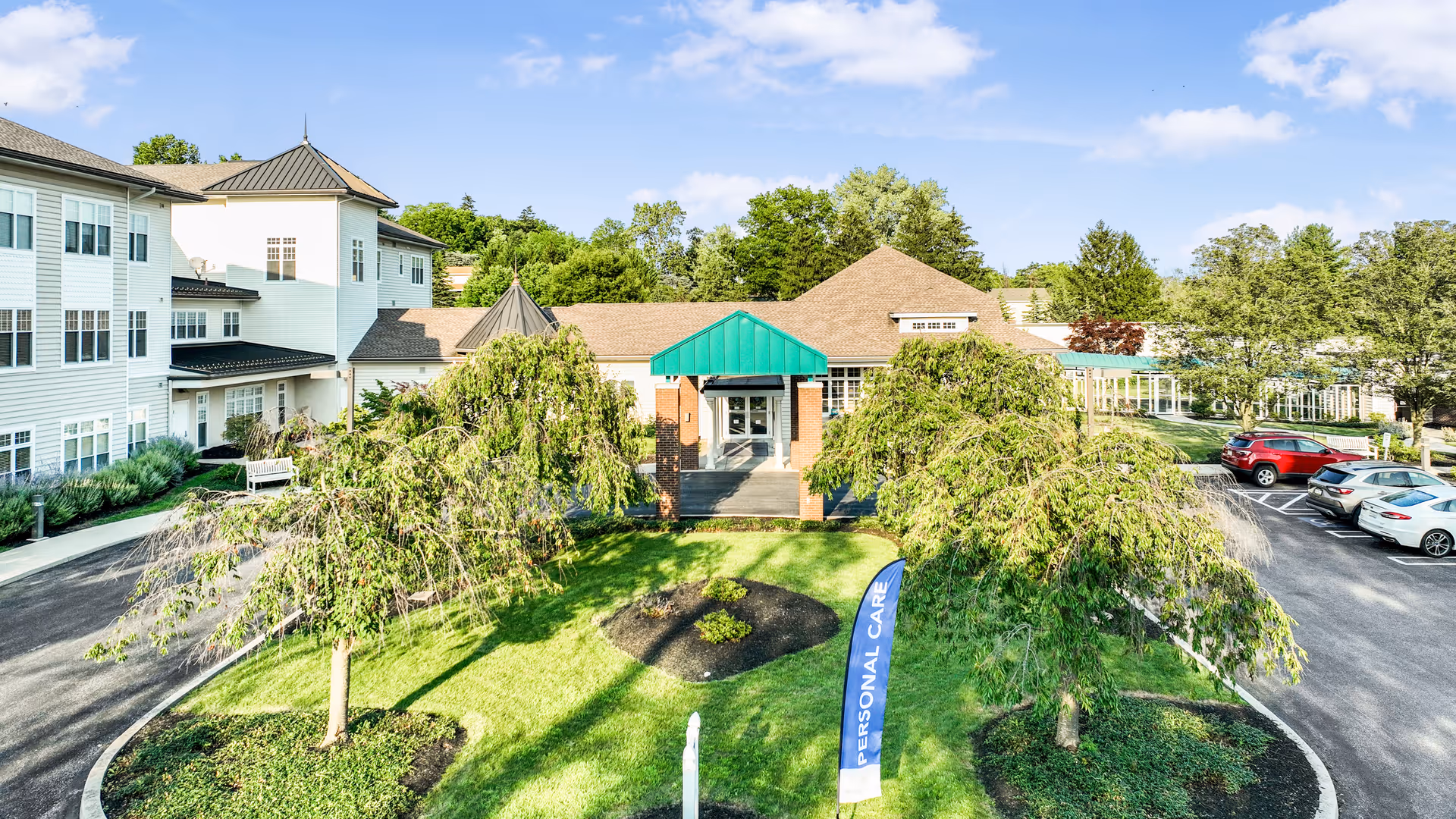Exterior front entrance of a senior living facility with a green awning, trees, landscaped lawn, and a parking lot.