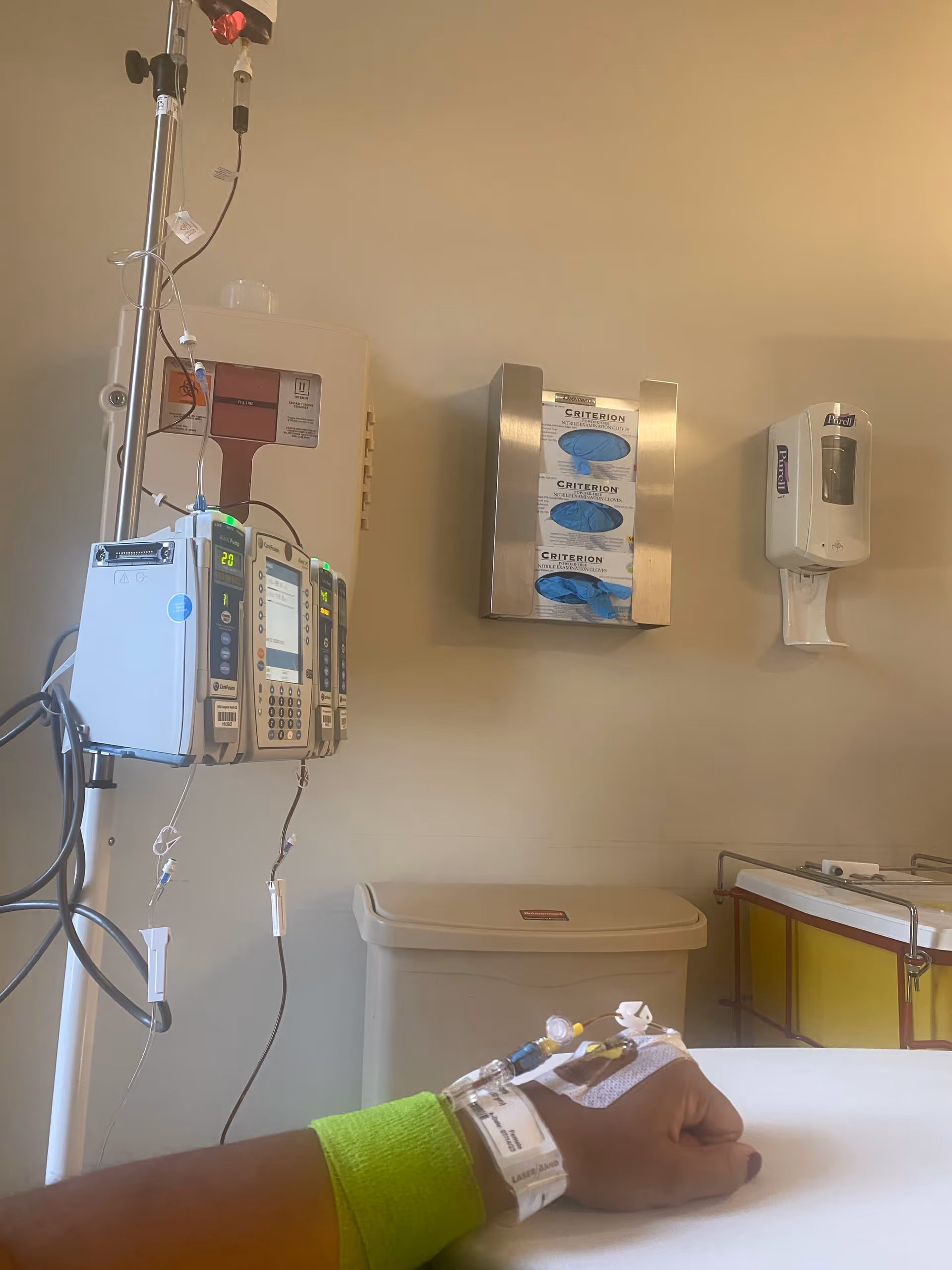 Patient arm with IV infusion and medical monitors in a hospital room.
