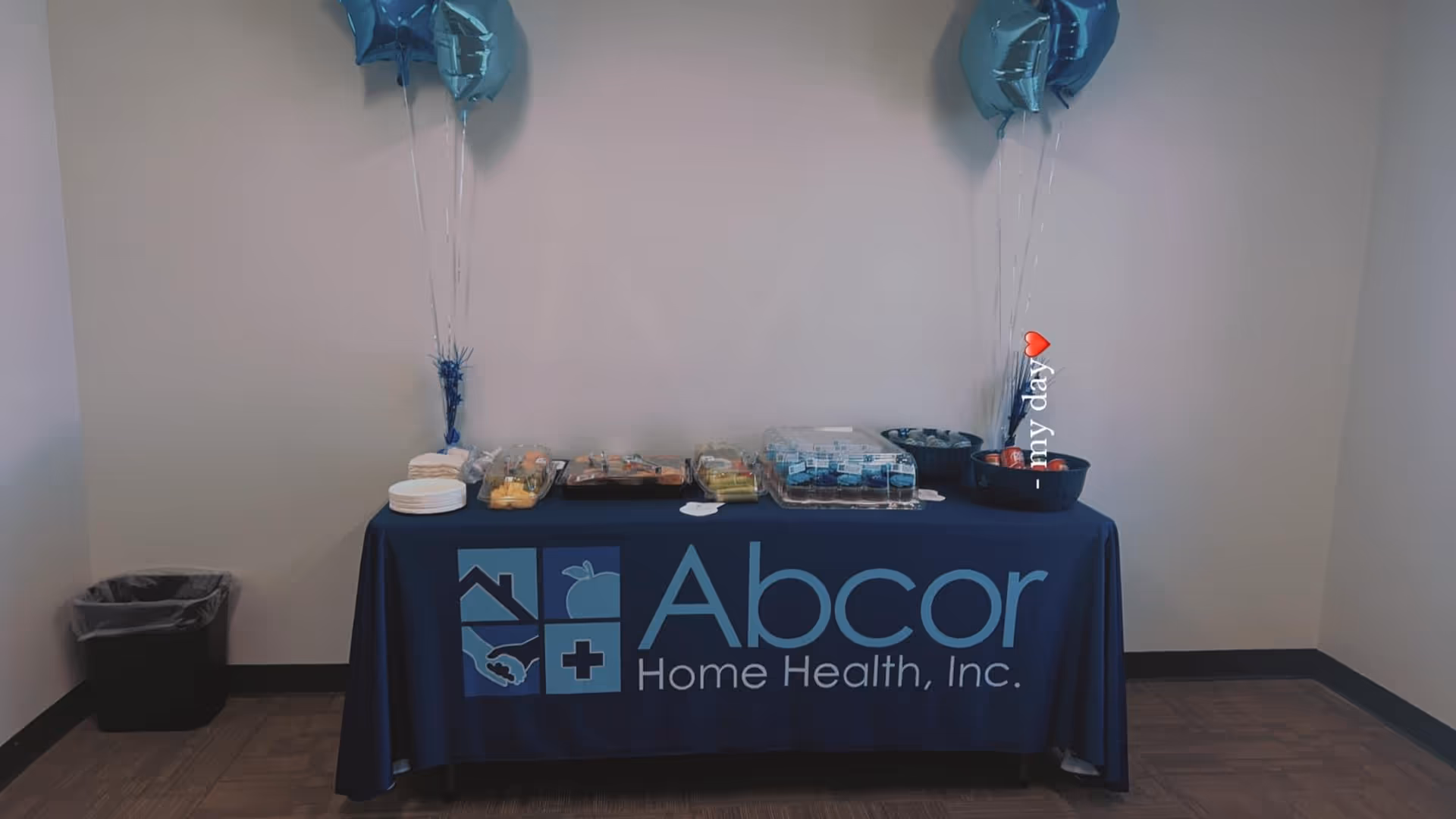 Indoor reception table with Abcor Home Health, Inc. tablecloth, snacks, and balloons.