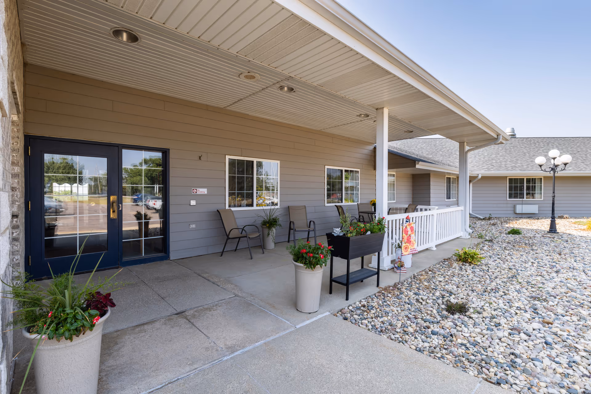 Covered front entrance and porch of a senior living building with seating, potted plants, and a rock-covered yard.