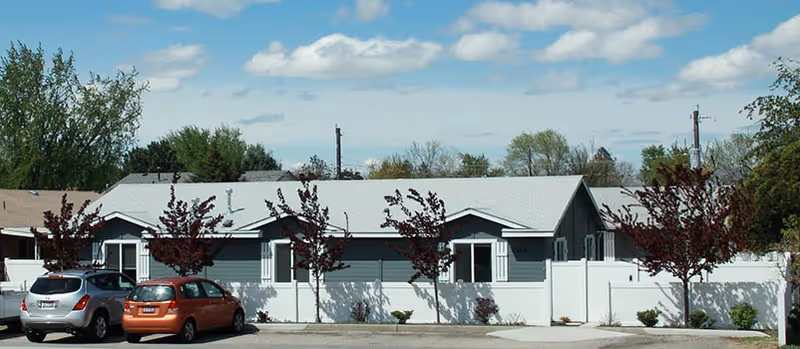 Single-story assisted living building with a white fence, small trees in front, and two parked cars under a partly cloudy sky.