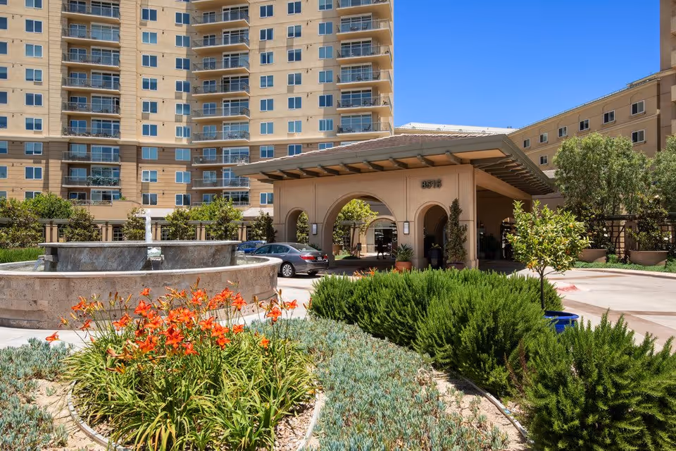 Exterior view of Vi at La Jolla Village, a CCRC, showing the main entrance with a covered drop-off area, a large multi-story building in the background, a circular water fountain, and landscaped garden with orange flowers and green shrubs under a clear blue sky.