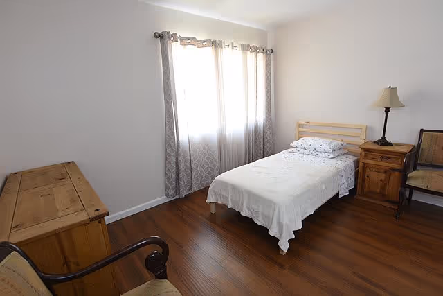 A simple bedroom with a single bed covered in white bedding, a wooden nightstand with a lamp, two chairs, a wooden dresser, and a window with patterned curtains letting in natural light.