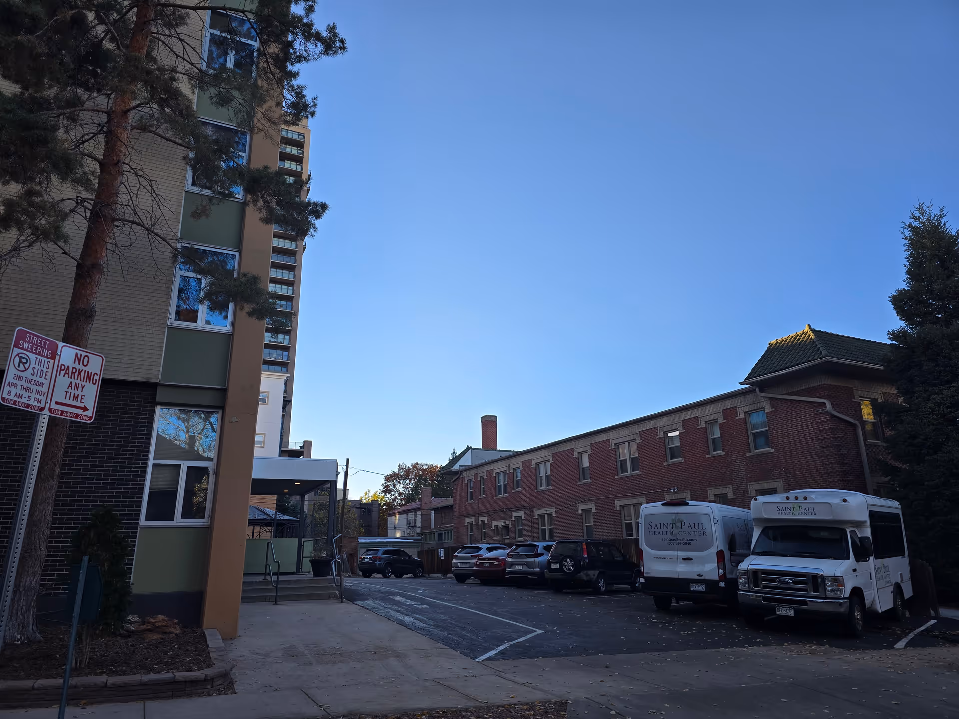 Parking lot and entrance area showing St Paul Health Center building with parked service vans and cars under a clear blue sky.