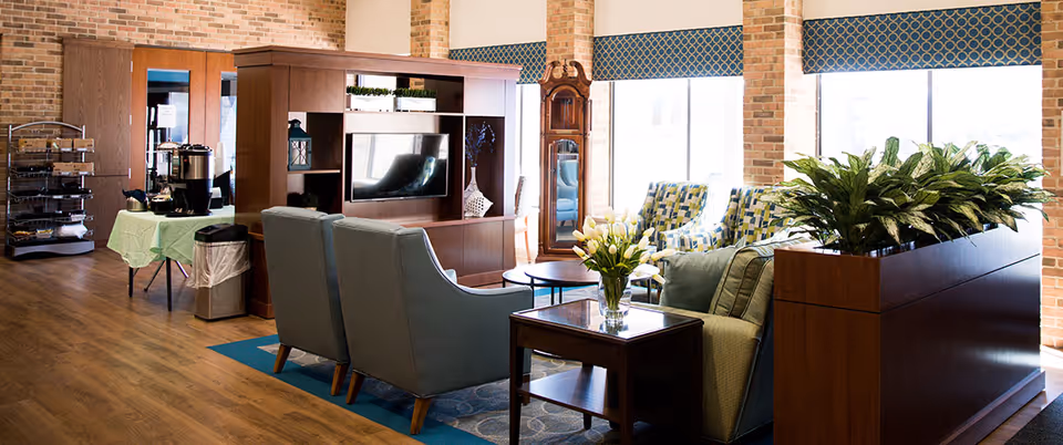 A bright and spacious senior living facility common area with wooden flooring, brick walls, and large windows with blue patterned valances. The room features comfortable seating including blue armchairs and a sofa around a wooden coffee table with a vase of white tulips. There is a wooden entertainment unit with a TV, a tall grandfather clock, and a plant divider. In the background, a table with a green tablecloth holds coffee and refreshments, and a metal rack with snacks is visible.