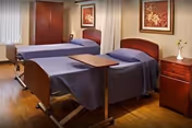 Two hospital-style beds with blue covers, wooden furniture, and framed wall art in a shared bedroom.
