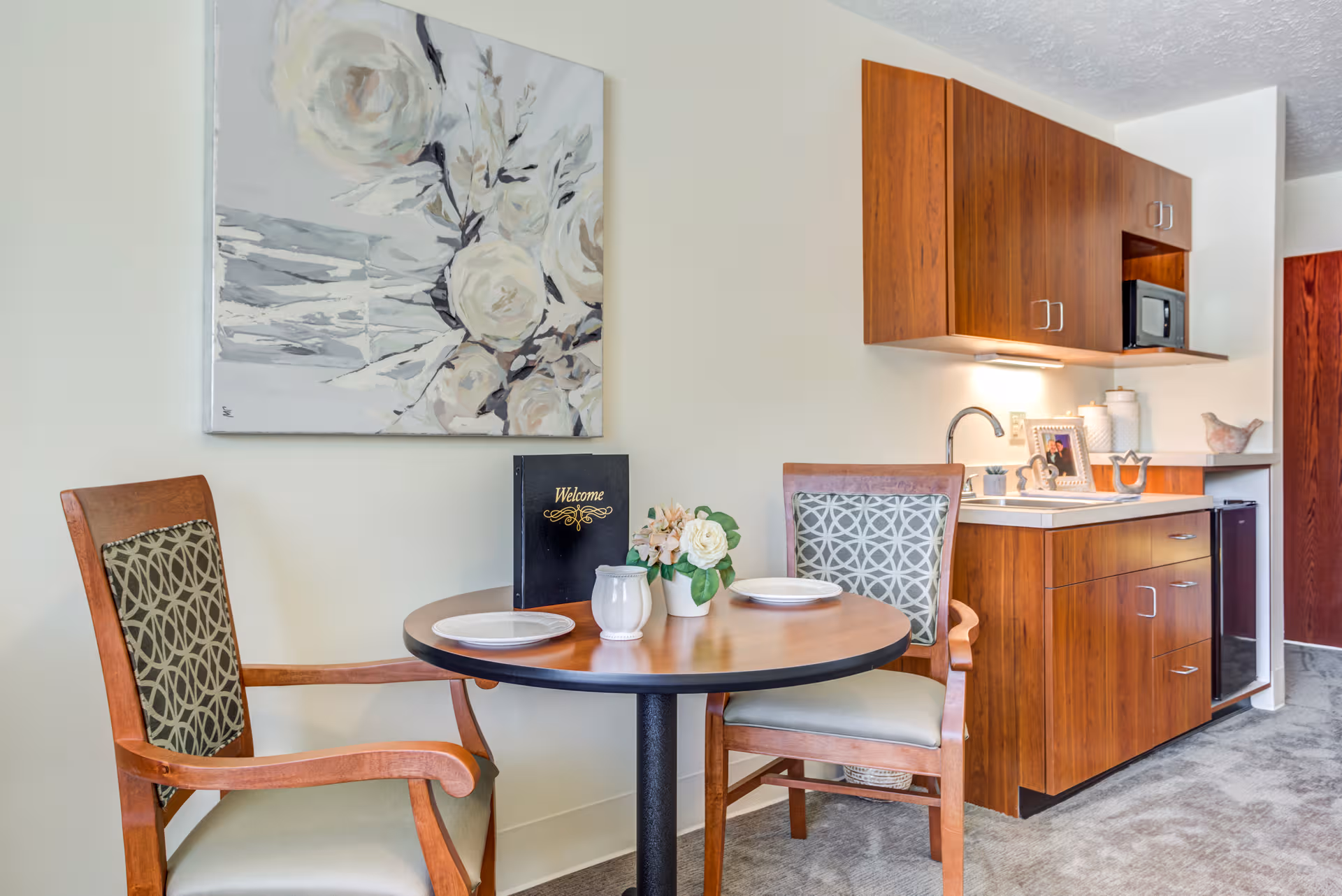 Round dining table with two chairs beside wood kitchen cabinets in a senior living suite.