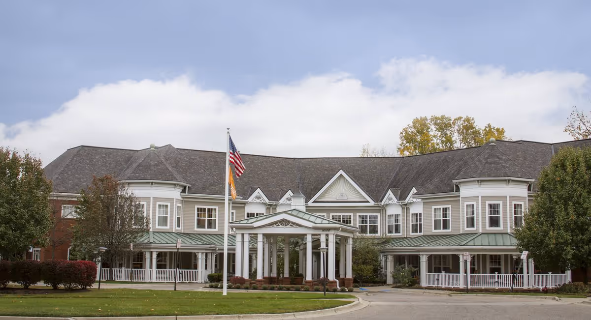 Front exterior view of a large, two-story senior living facility building with a covered entrance, multiple windows, and an American flag on a flagpole in front. The building is surrounded by trees and bushes under a partly cloudy sky.