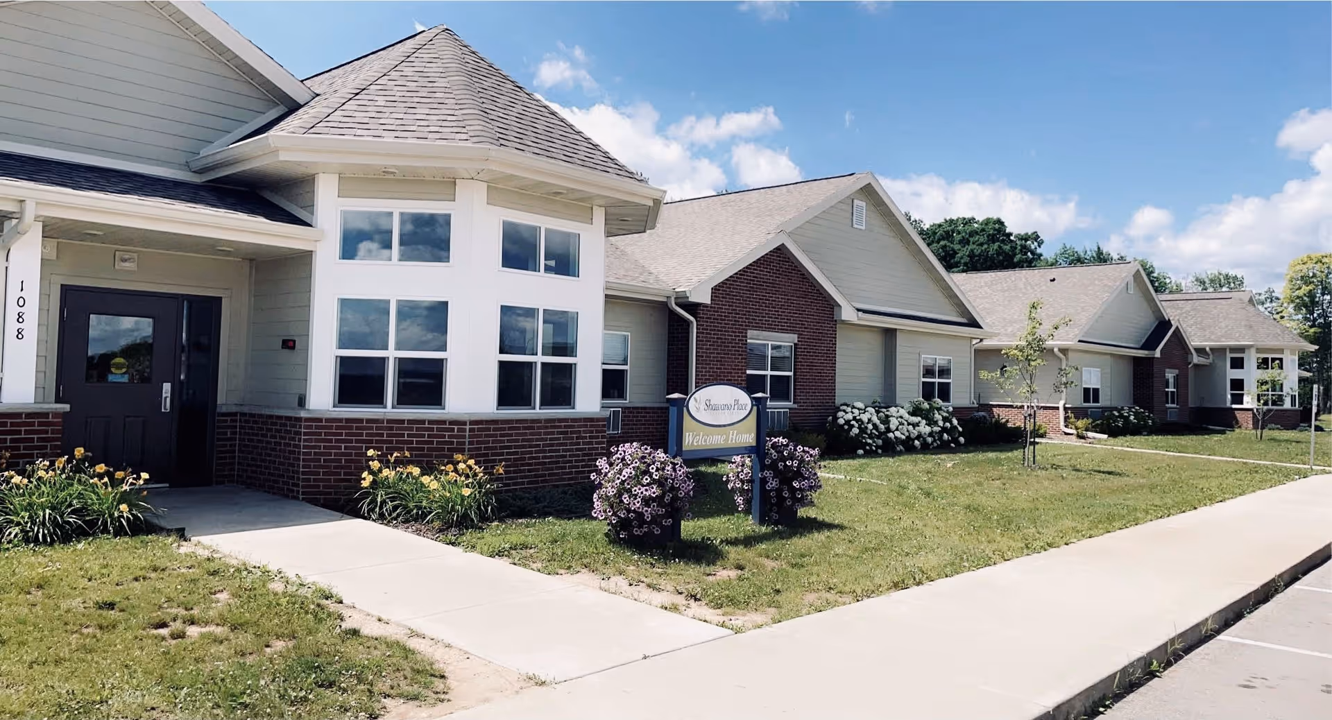 Exterior view of Shawano Place Senior Living building on a sunny day, showing a brick and siding facade with multiple windows, a main entrance door, a sidewalk, green lawn, flowers, and a sign that reads 'Shawano Place Welcome Home'.