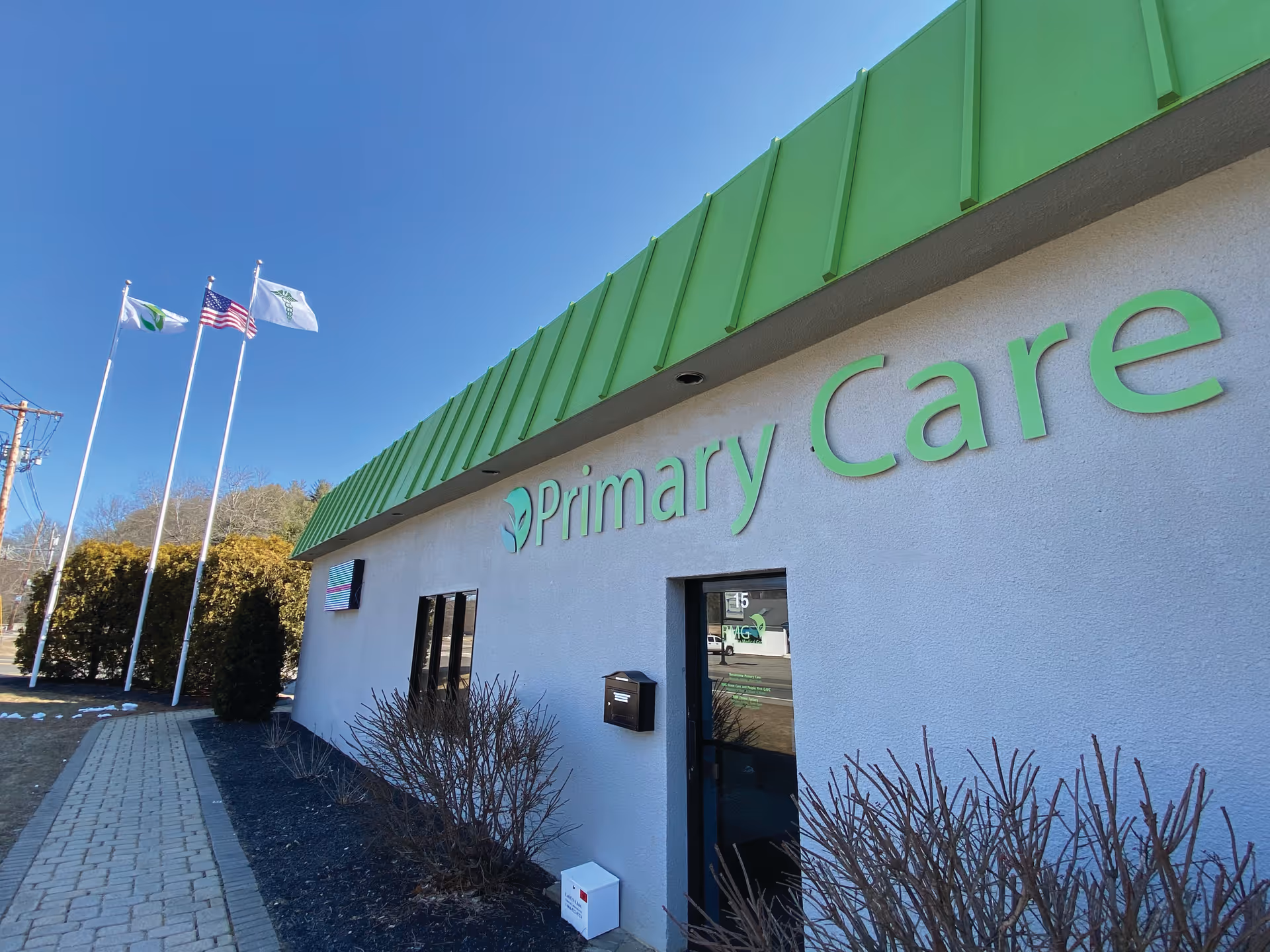White building with green roof and Primary Care sign, flags outside, and a sidewalk.