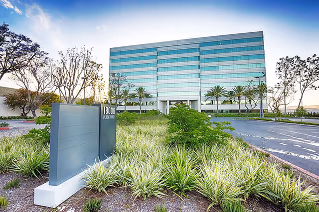 Modern glass-front building with palm trees and a manicured entrance landscape.