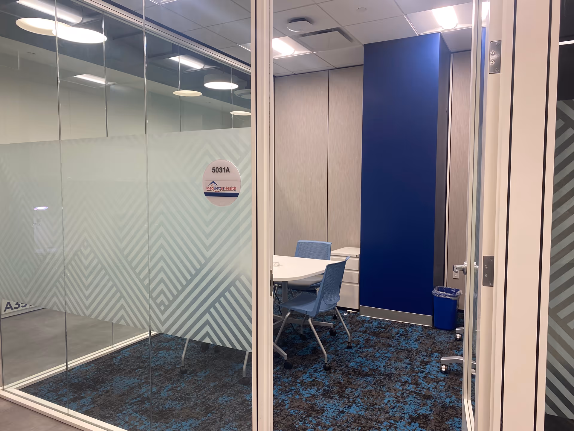 Glass-walled conference room with blue accents, table and blue chairs inside a modern facility.