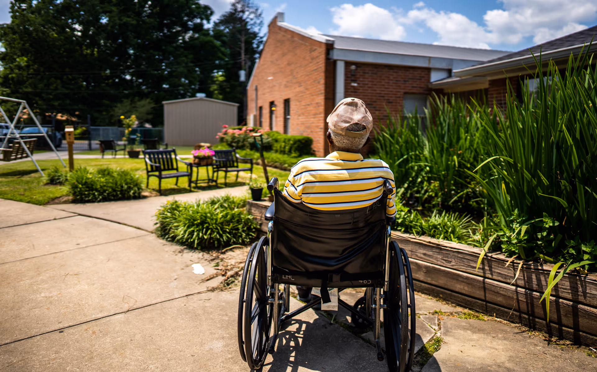 A person in a wheelchair wearing a yellow and white striped shirt and a beige cap is outdoors on a paved pathway. The scene includes green plants, a brick building, outdoor chairs, a table with flowers, and a swing set in the background under a partly cloudy sky.