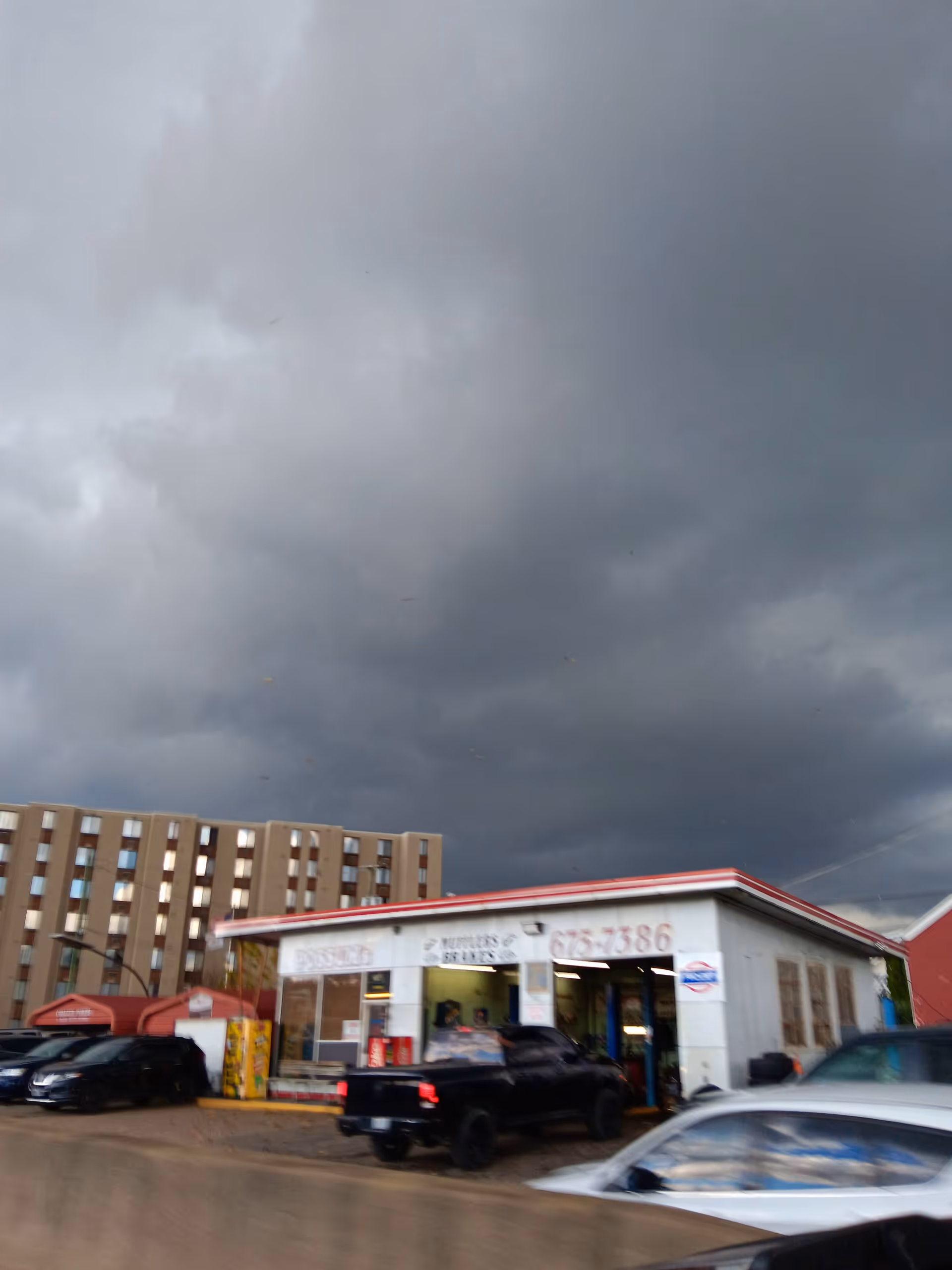 Stormy dark clouds loom over a small white storefront with parked cars.