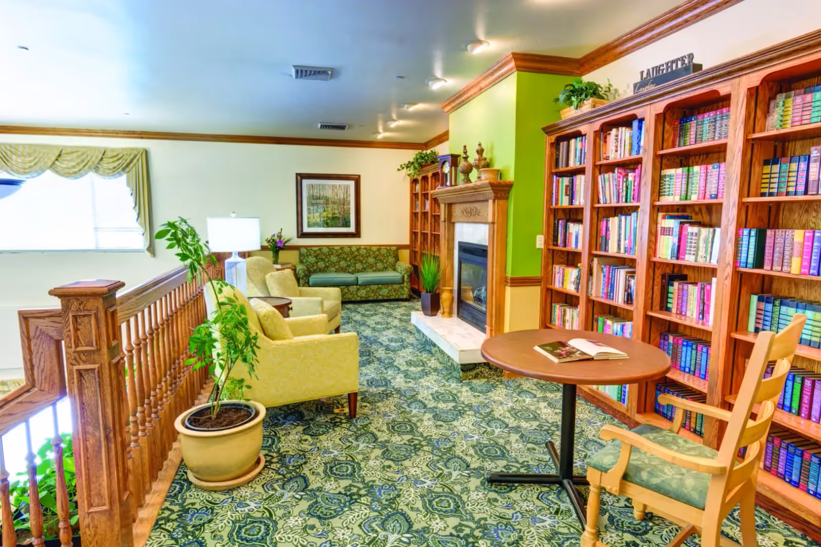 A bright senior living lounge with armchairs, a fireplace, bookshelves, and a small table overlooking a staircase.