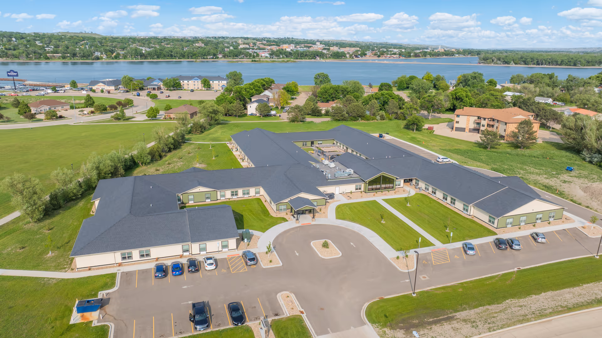 Aerial view of Peaceful Pines Senior Living campus by a river, with curved drive and parking.
