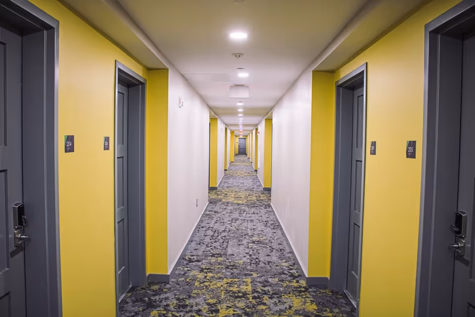 A long, well-lit hallway in a senior living facility with yellow walls and gray doors on both sides, each door labeled with room numbers such as 204 and 205. The floor is covered with a patterned carpet in shades of gray and yellow.