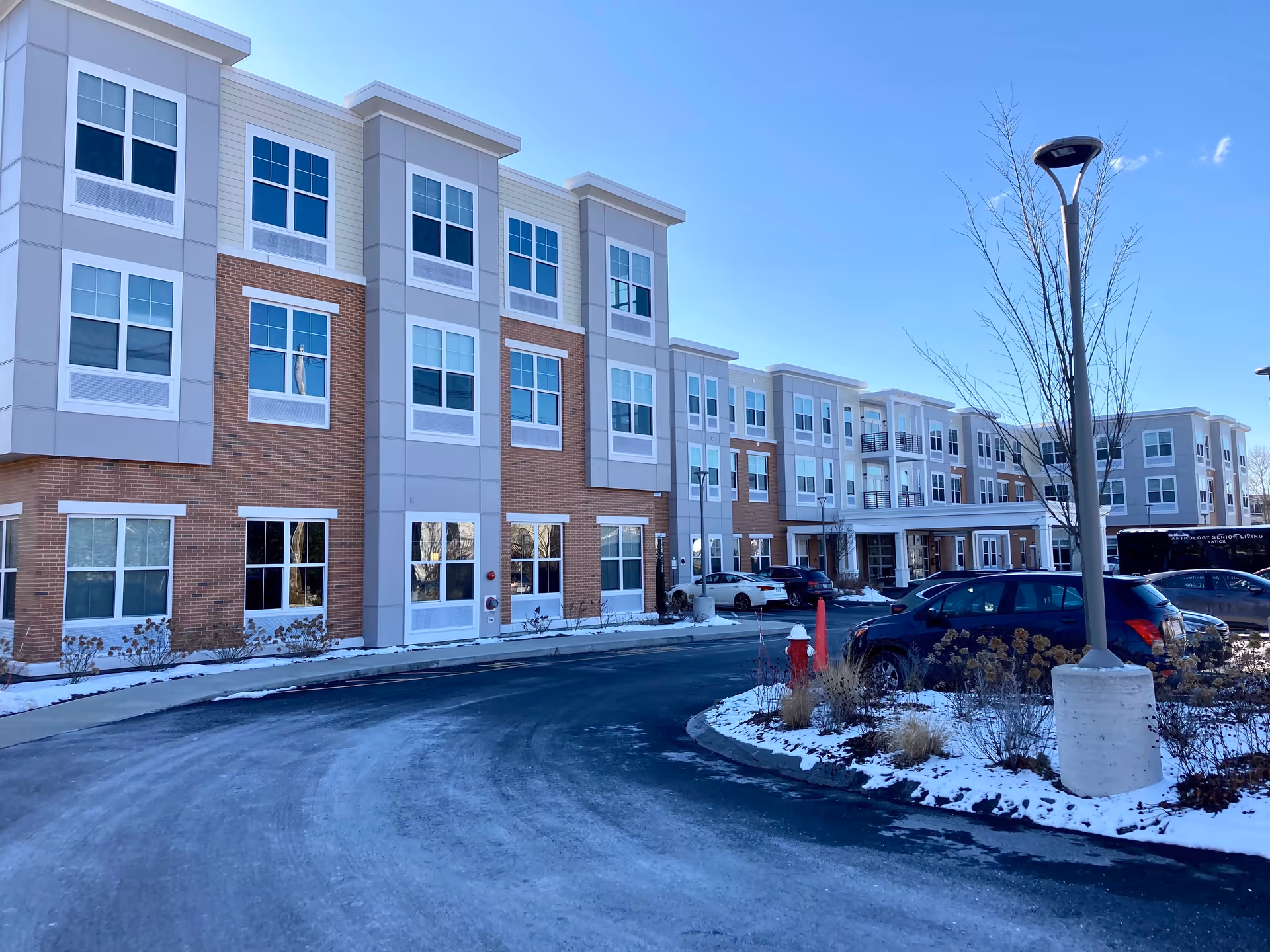 Exterior front of a three-story residential building with a curved driveway, parked cars, and light snow.
