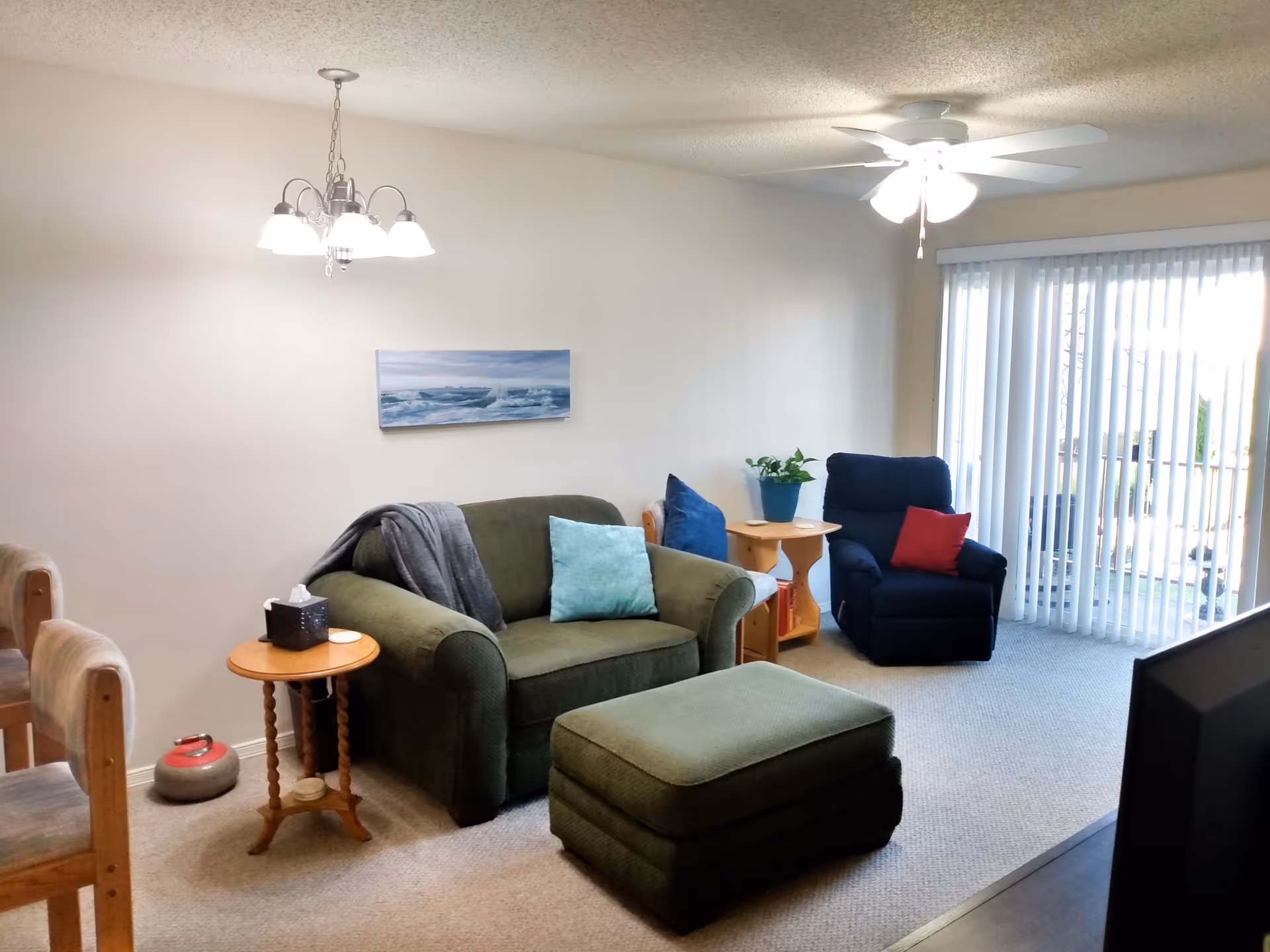 Cozy living room with a green loveseat and ottoman, blue recliner, side tables, ceiling fan, and sliding glass doors with vertical blinds.