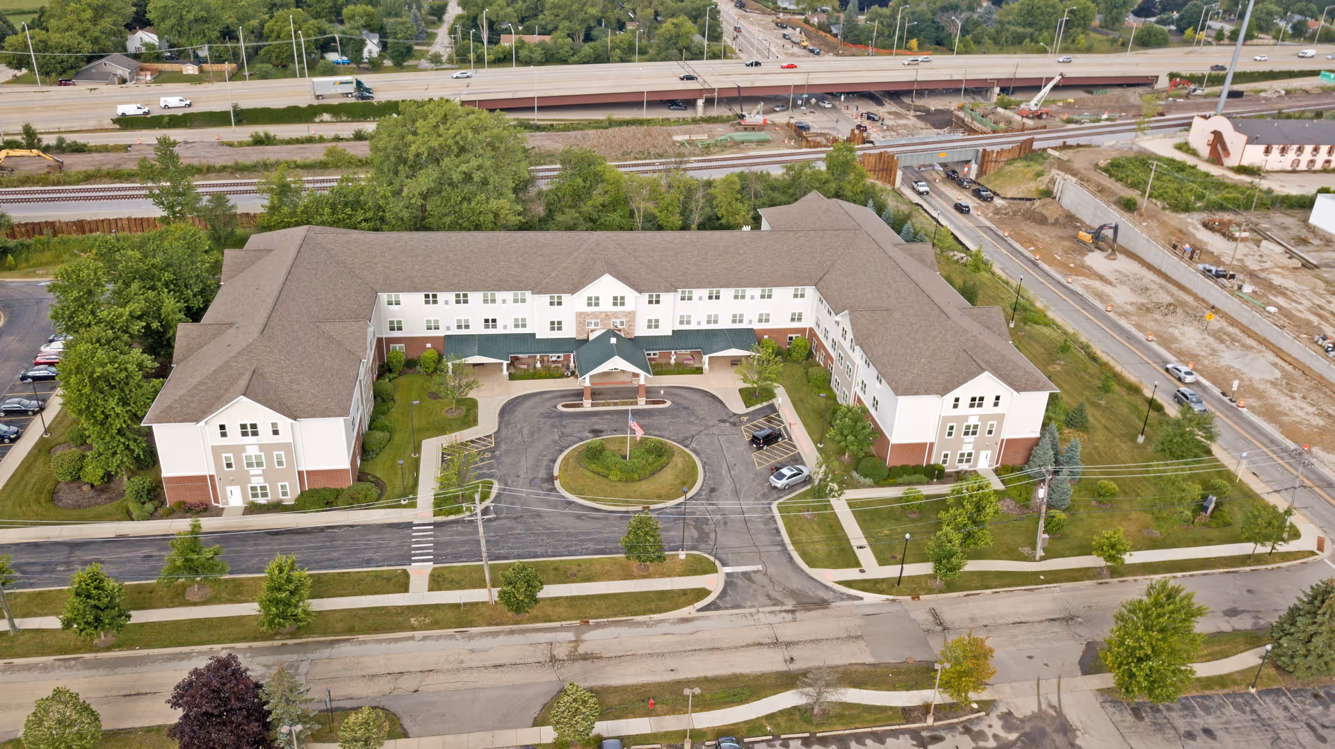 Aerial view of a large, U-shaped senior living facility building with a circular driveway and landscaped greenery in front. The building has three stories with a mix of white and brick exterior walls and a green roof over the entrance. Surrounding the facility are roads, parking areas, trees, and a nearby highway with vehicles.