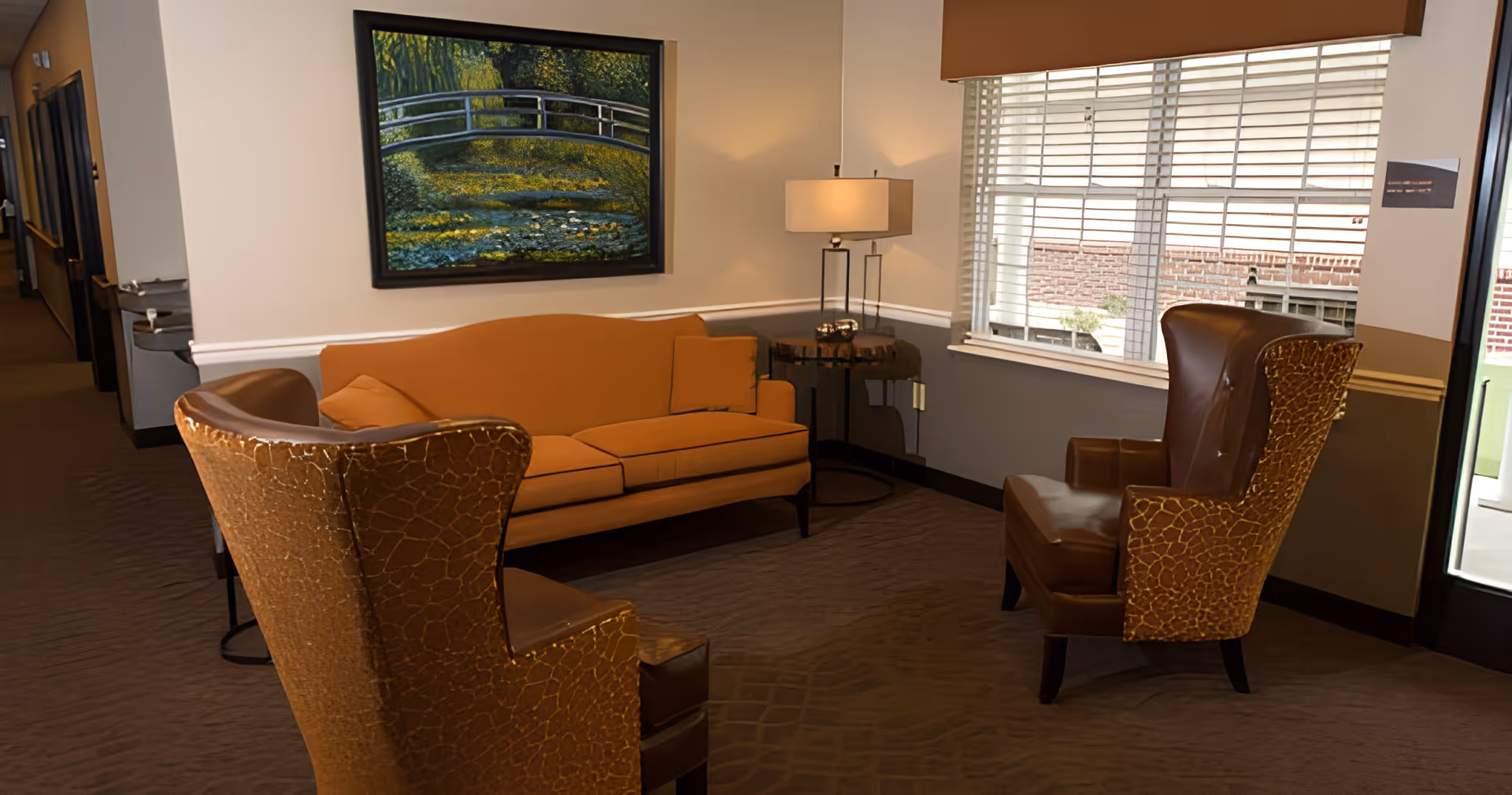 A cozy sitting area in a senior living facility with an orange sofa, two brown leather armchairs with textured backs, a side table with a lamp, a large window with blinds, and a painting of a bridge over a stream on the wall.