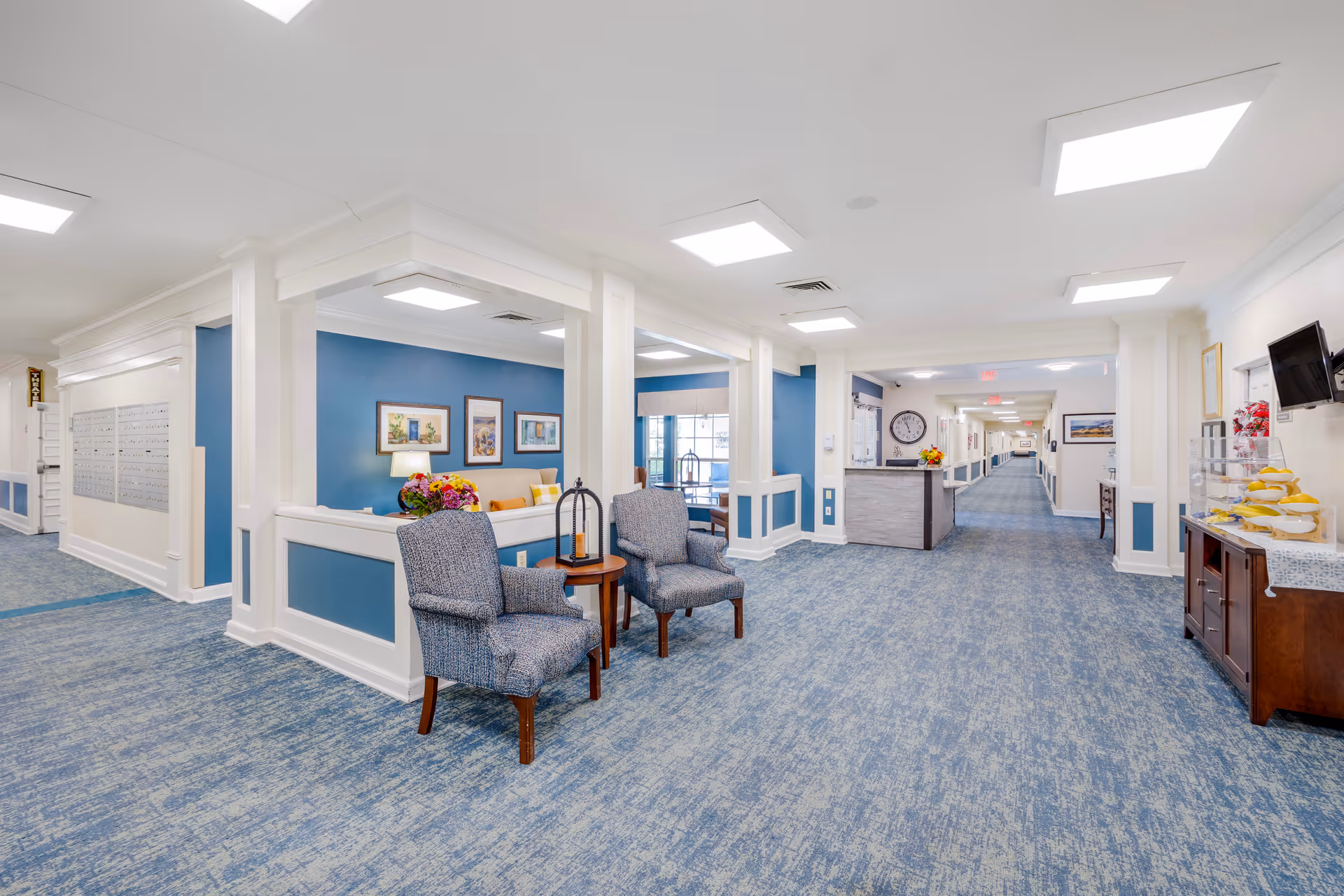 A bright and spacious senior living facility hallway with blue patterned carpet and white walls accented with blue panels. Two upholstered chairs with a small round table and decorative lantern are positioned near an open seating area with a sofa, colorful cushions, and framed artwork on a blue wall. The hallway extends into the distance with a reception desk, clock, and additional seating areas visible.