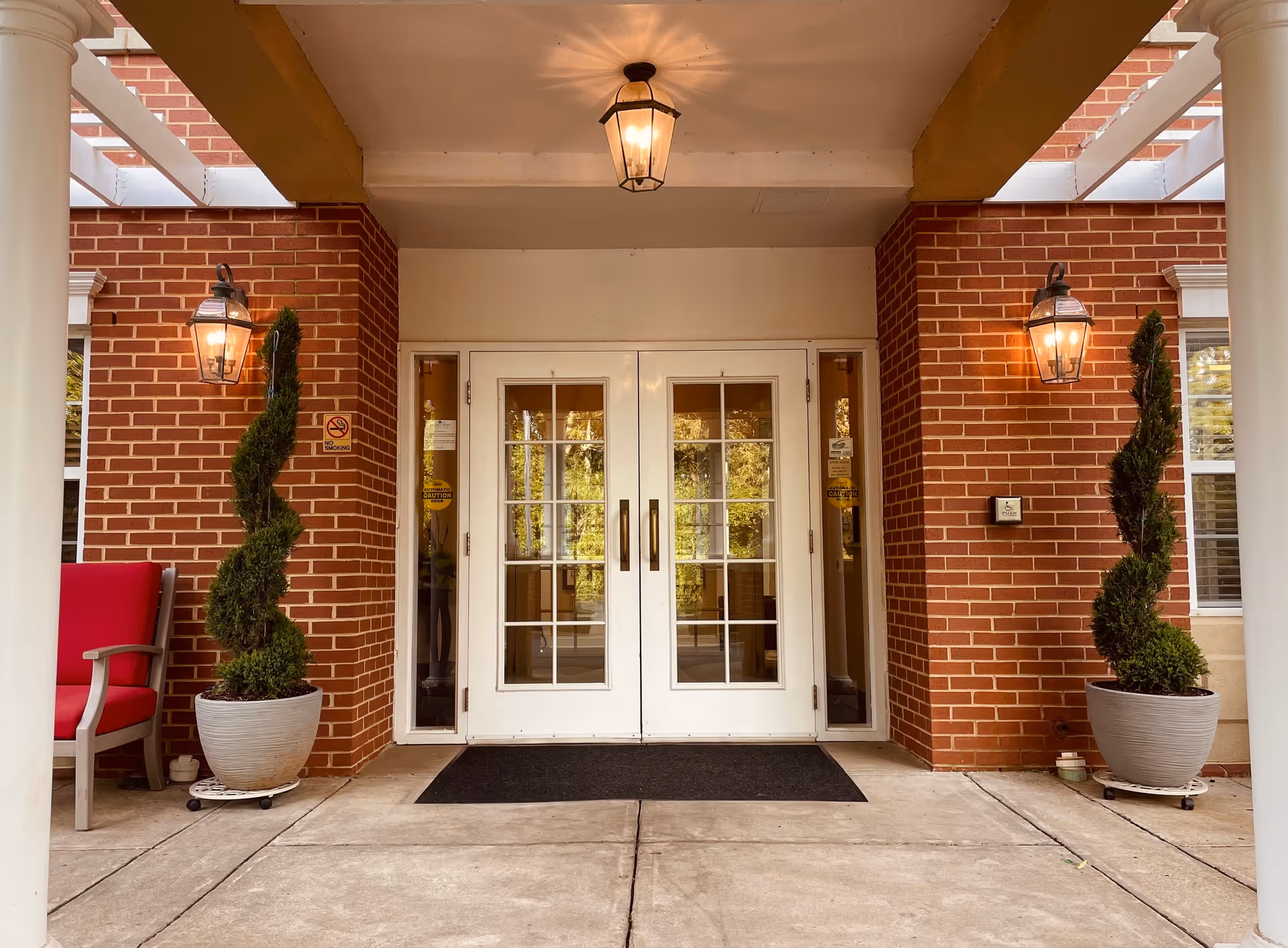 Entrance to a building with double white doors featuring glass panes, flanked by two spiral-shaped potted plants on either side. The entrance is framed by red brick walls, two wall-mounted lantern-style lights, a ceiling light fixture, and a red cushioned chair on the left side.