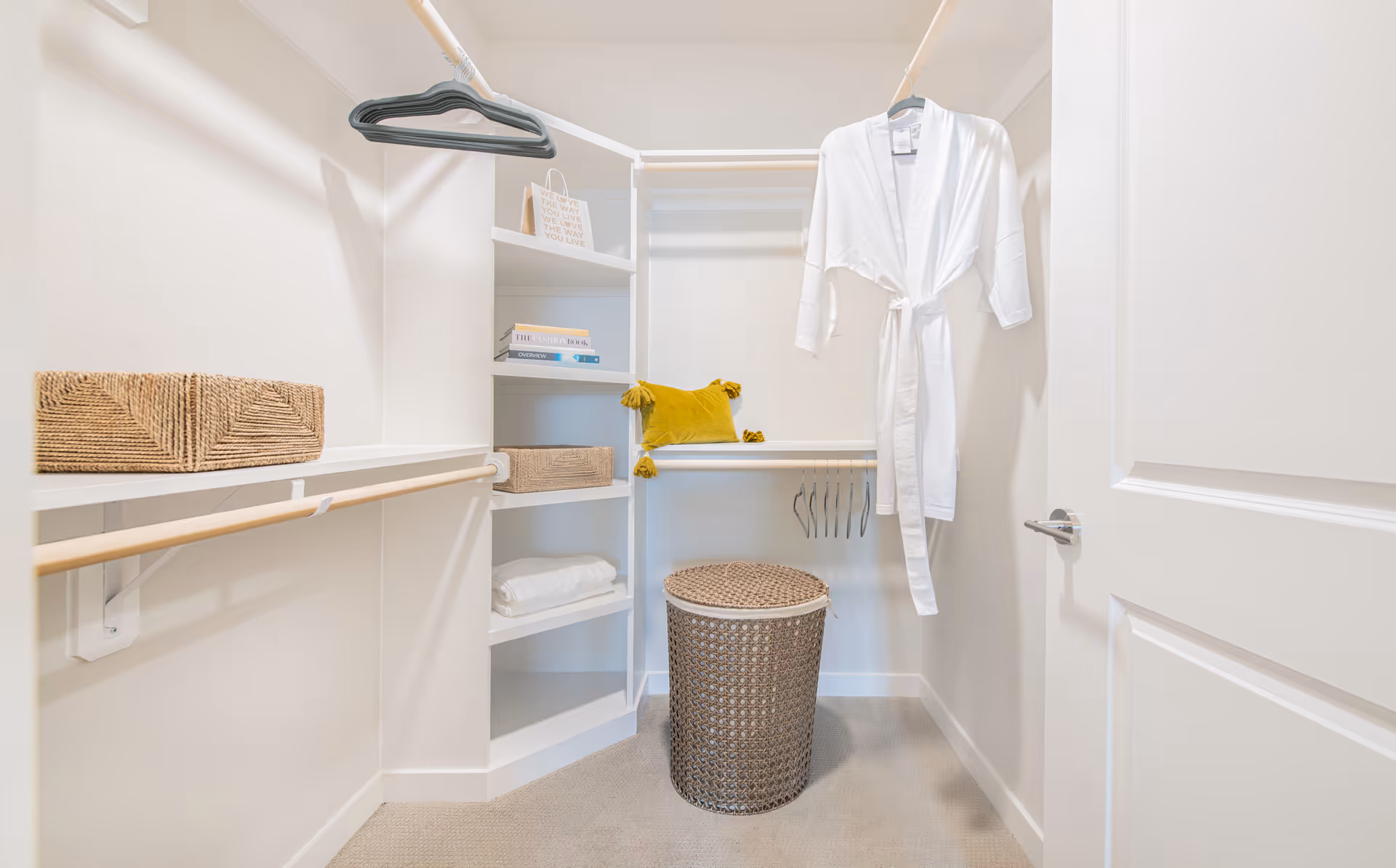 Well-lit walk-in closet with shelves, hanging rods, woven baskets, a white robe, and a wicker laundry hamper.