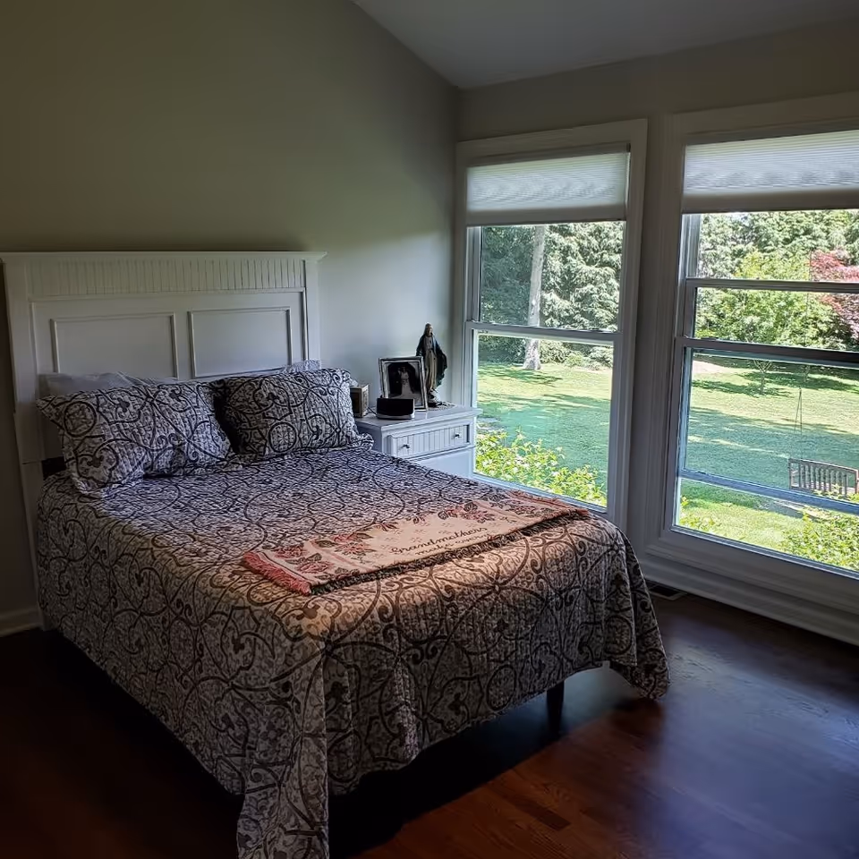 Bright bedroom with white headboard, patterned quilt, and large windows overlooking a green lawn.
