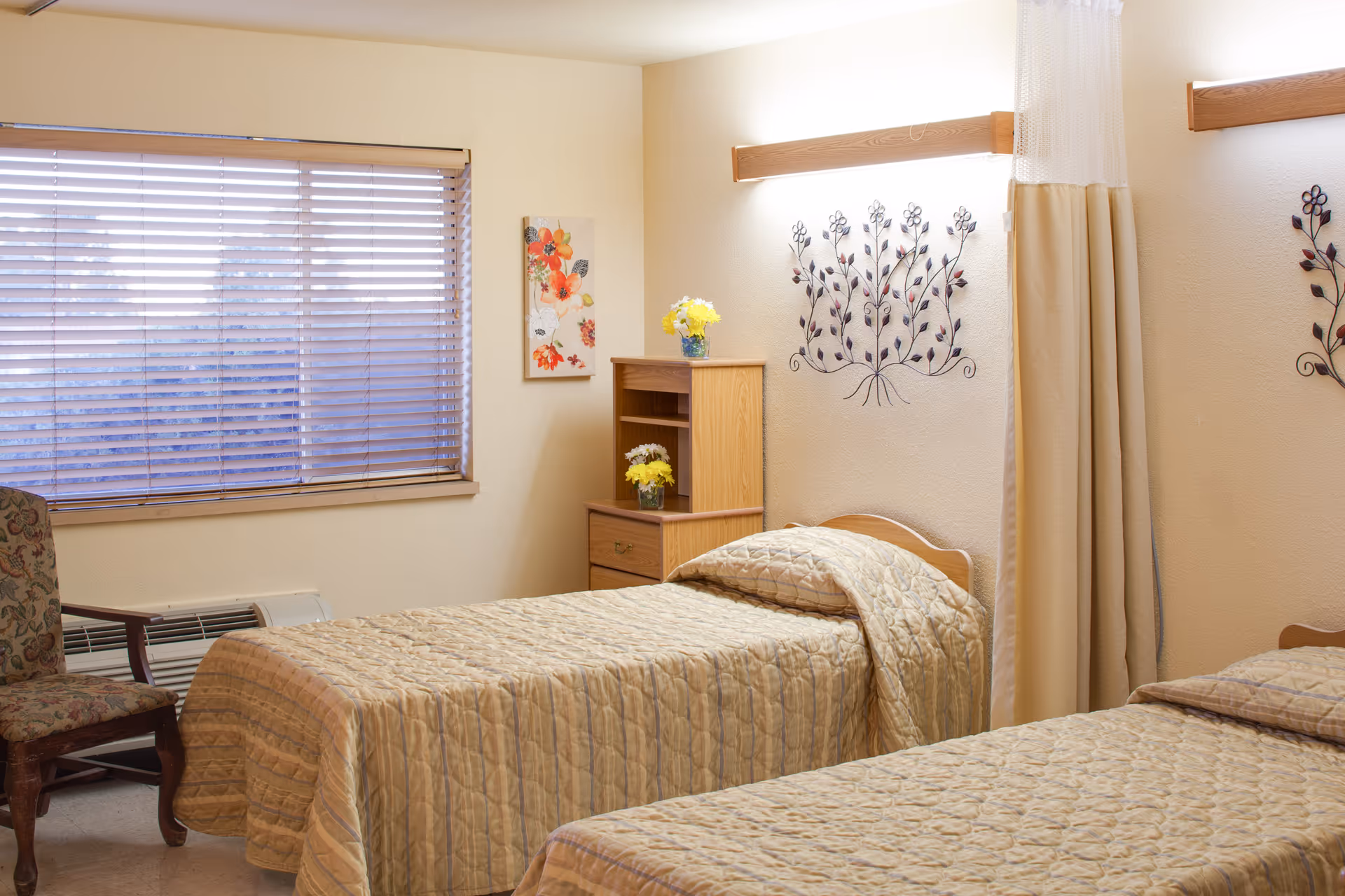 A cozy bedroom with two single beds covered in beige quilted bedspreads. There is a wooden nightstand with yellow flowers in a vase between the beds. A floral painting hangs on the wall next to a window with closed blinds. A cushioned chair with a floral pattern is placed near the window. The room has soft lighting and a privacy curtain partially visible on the right side.