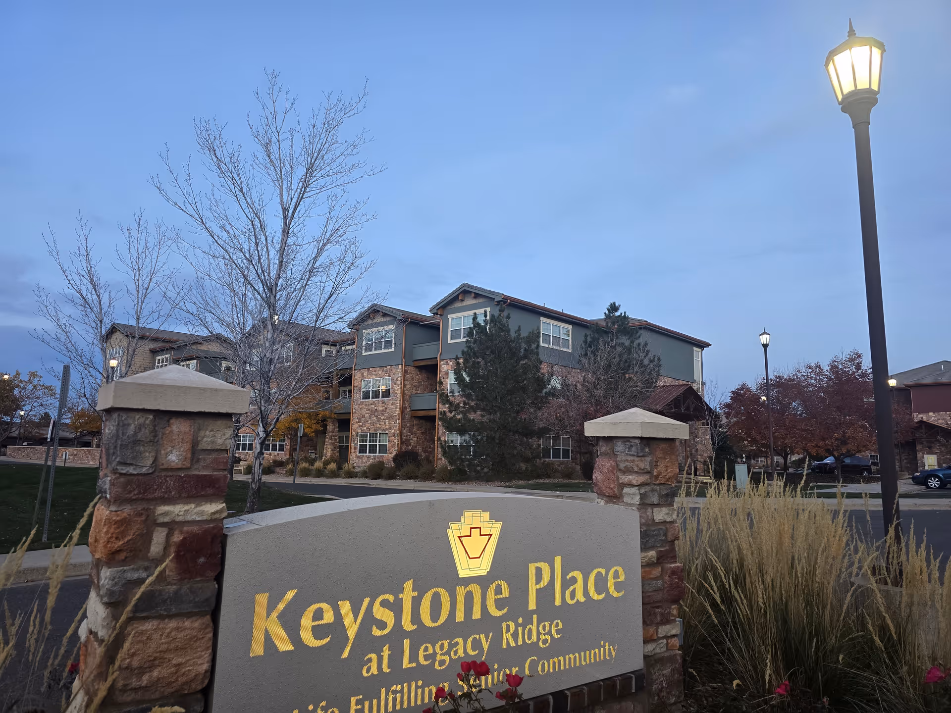Stone entrance sign reading "Keystone Place at Legacy Ridge" in front of a multi-story assisted living building with lamp posts and landscaping at dusk.